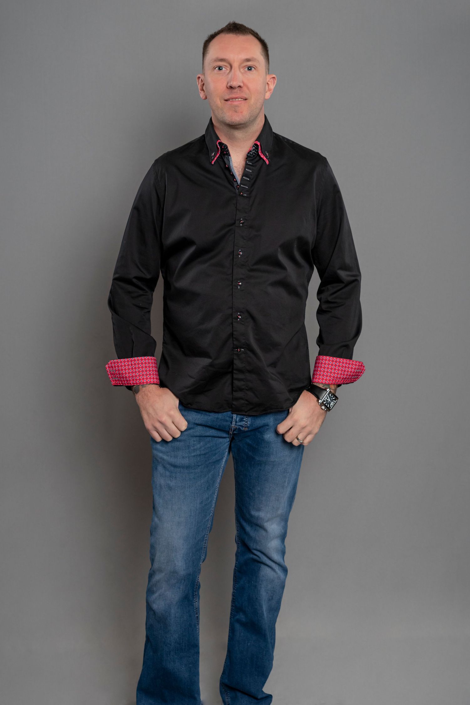 Professional portrait of man in black button-down shirt and jeans against grey background.