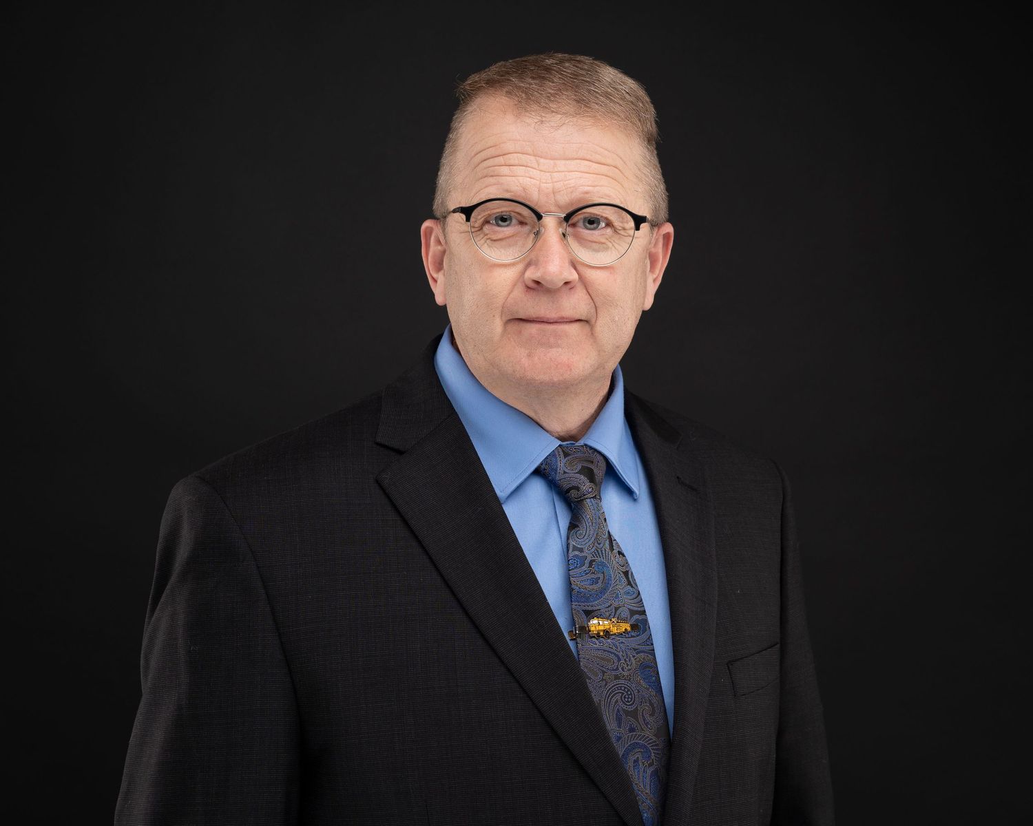 Professional portrait of person in dark suit and blue dress shirt with patterned tie against black background.