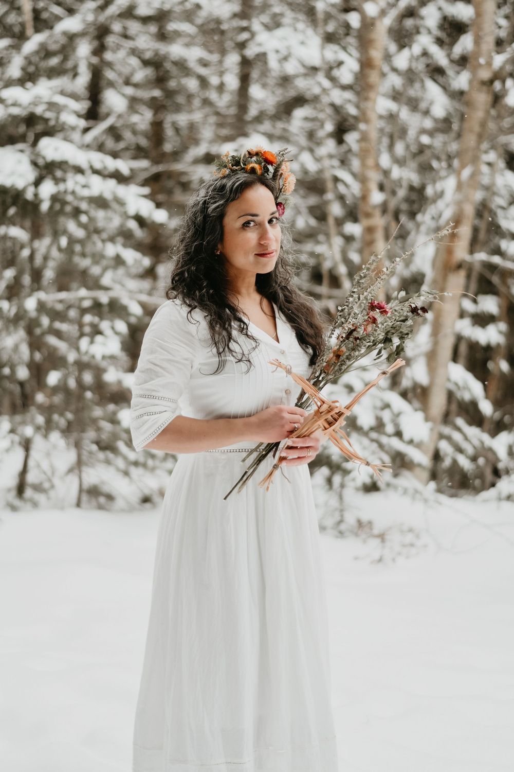 Person in white dress holds dried flowers in snowy winter forest.