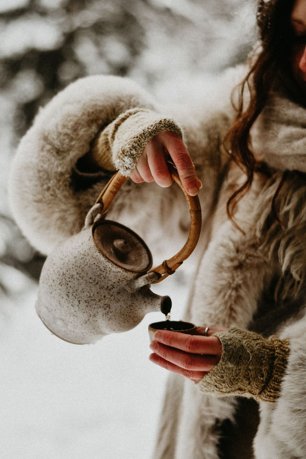 Hands in knit gloves pour hot drink from wooden teapot in winter setting.