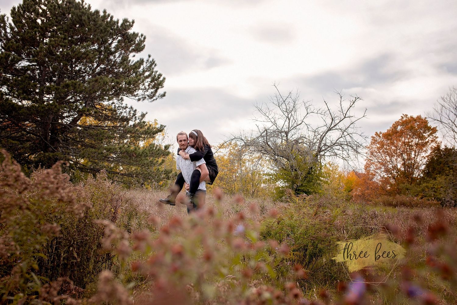 Couple embracing in autumn field with wildflowers and trees in background.
