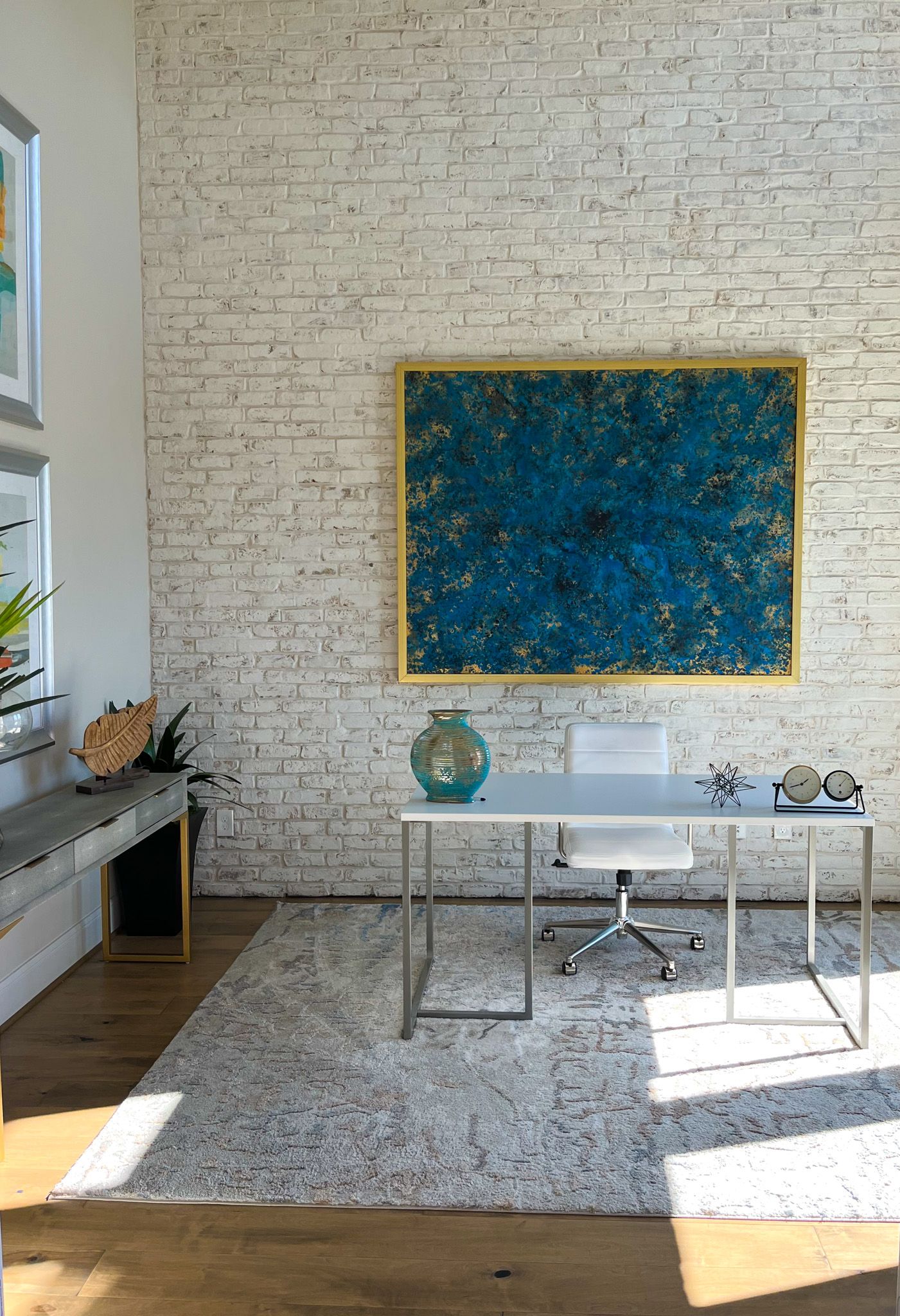Modern office space with white brick walls, blue abstract artwork, and a minimalist desk setup in natural sunlight.