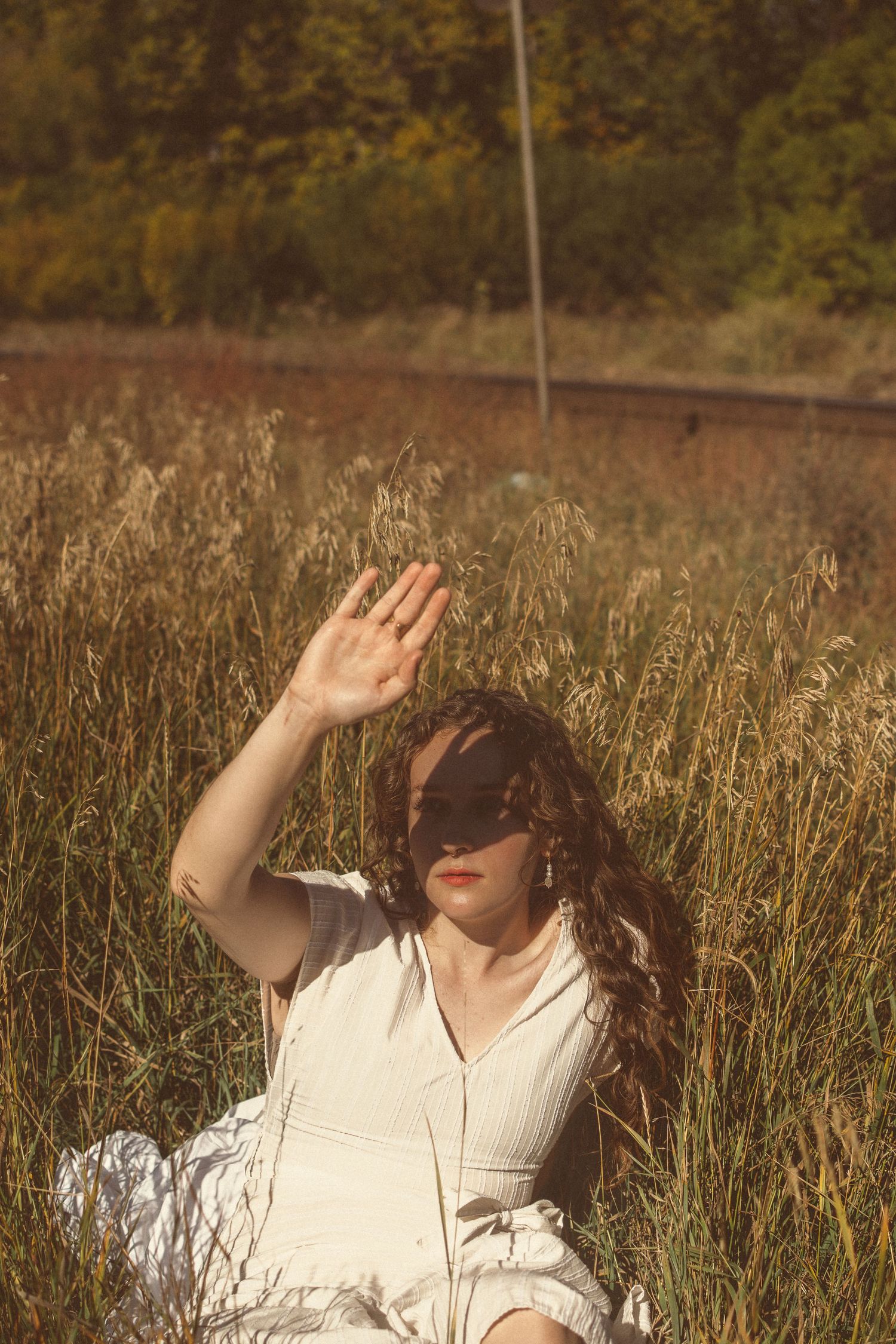 Someone in white dress raises their hand while sitting in tall grass by railroad tracks.