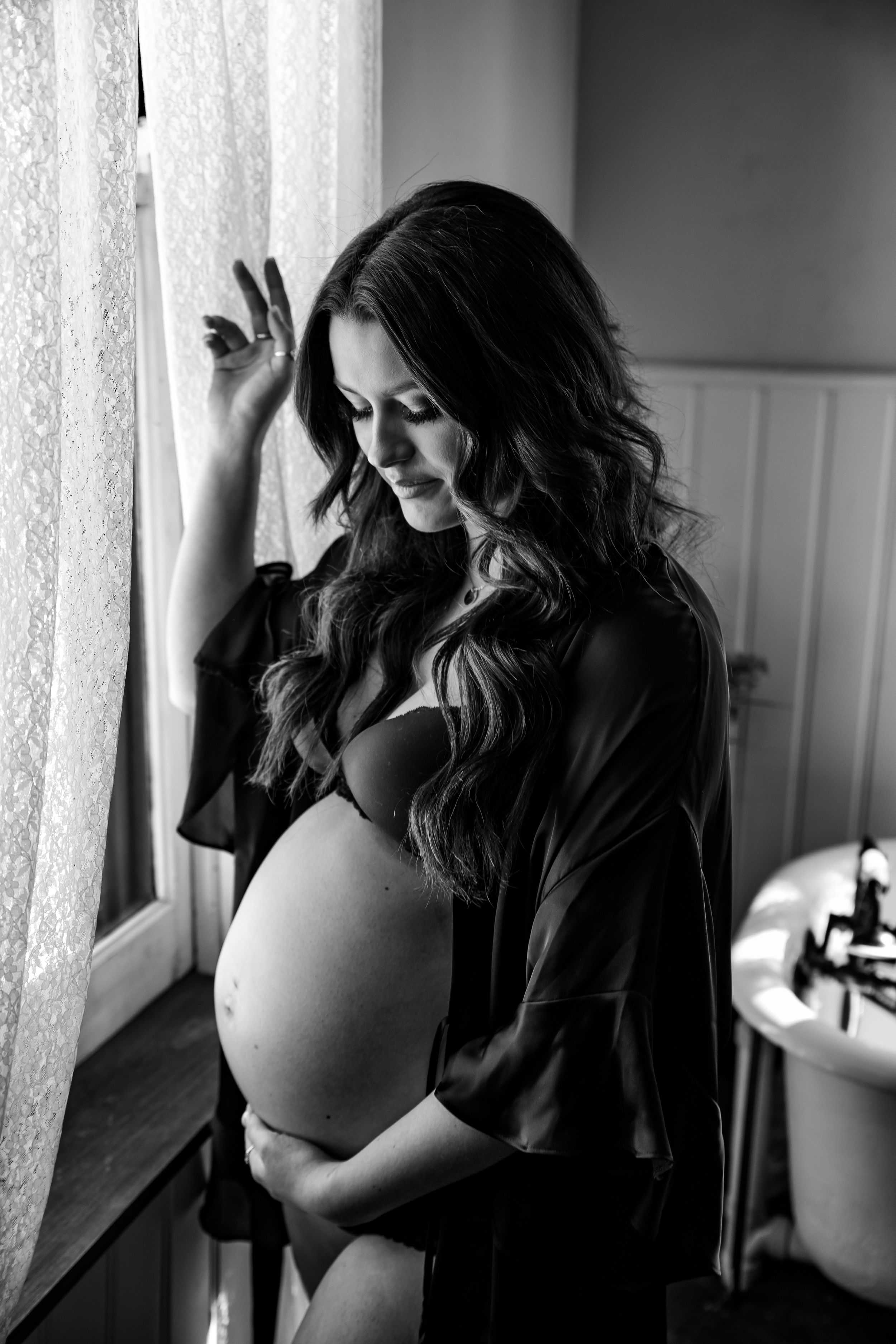 A silhouette of an expectant mother stands by a window in a dramatic black and white portrait.