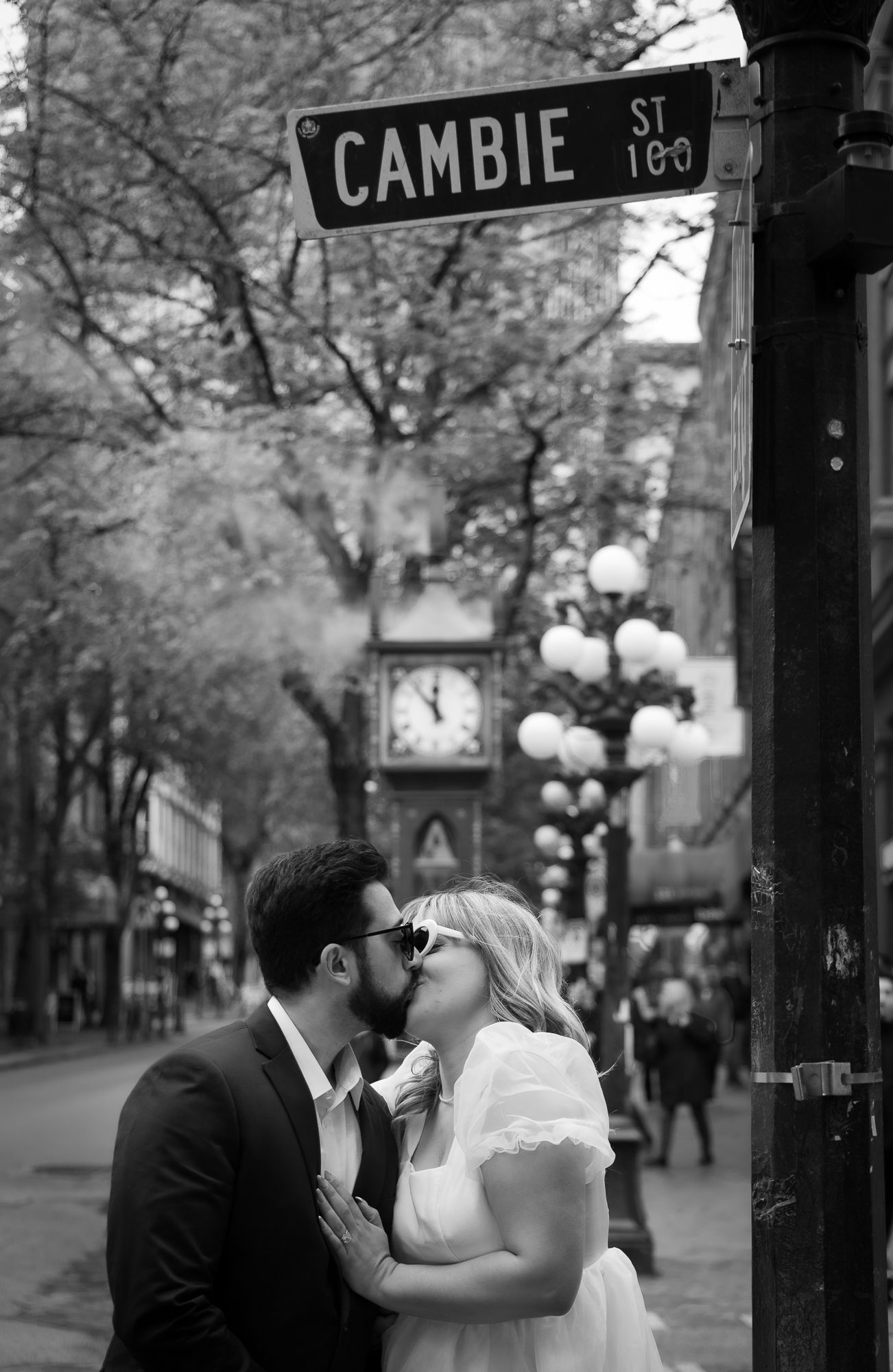 A couple kisses in Gastown, wearing fun sunglasses, under the Cambie St sign.