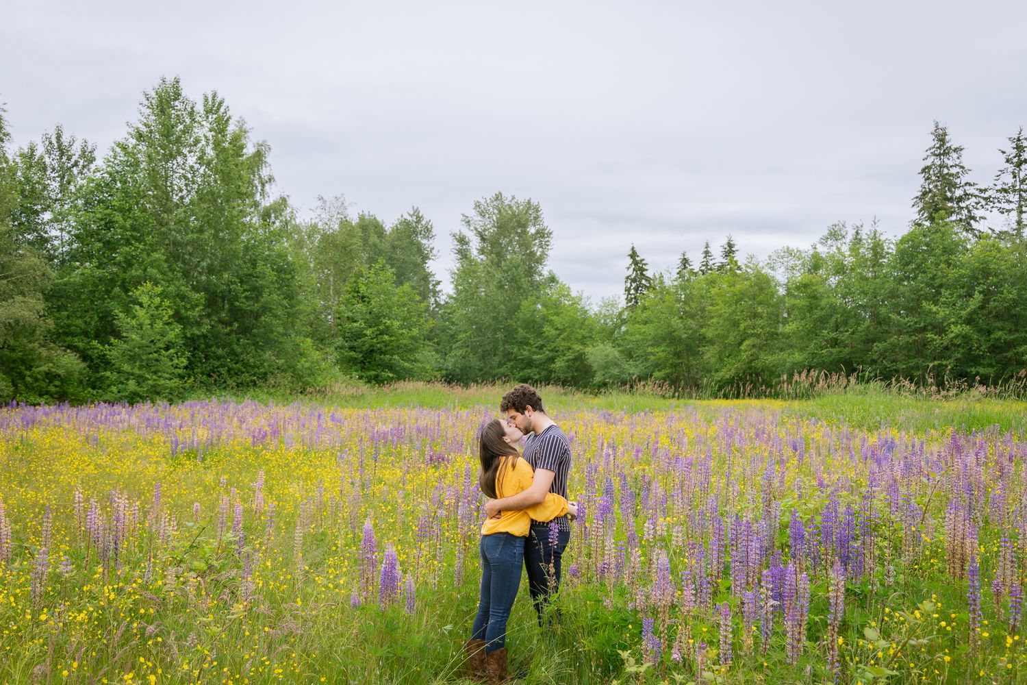 A young man and young woman kiss while holding each other in a bright green field of purple flowers.