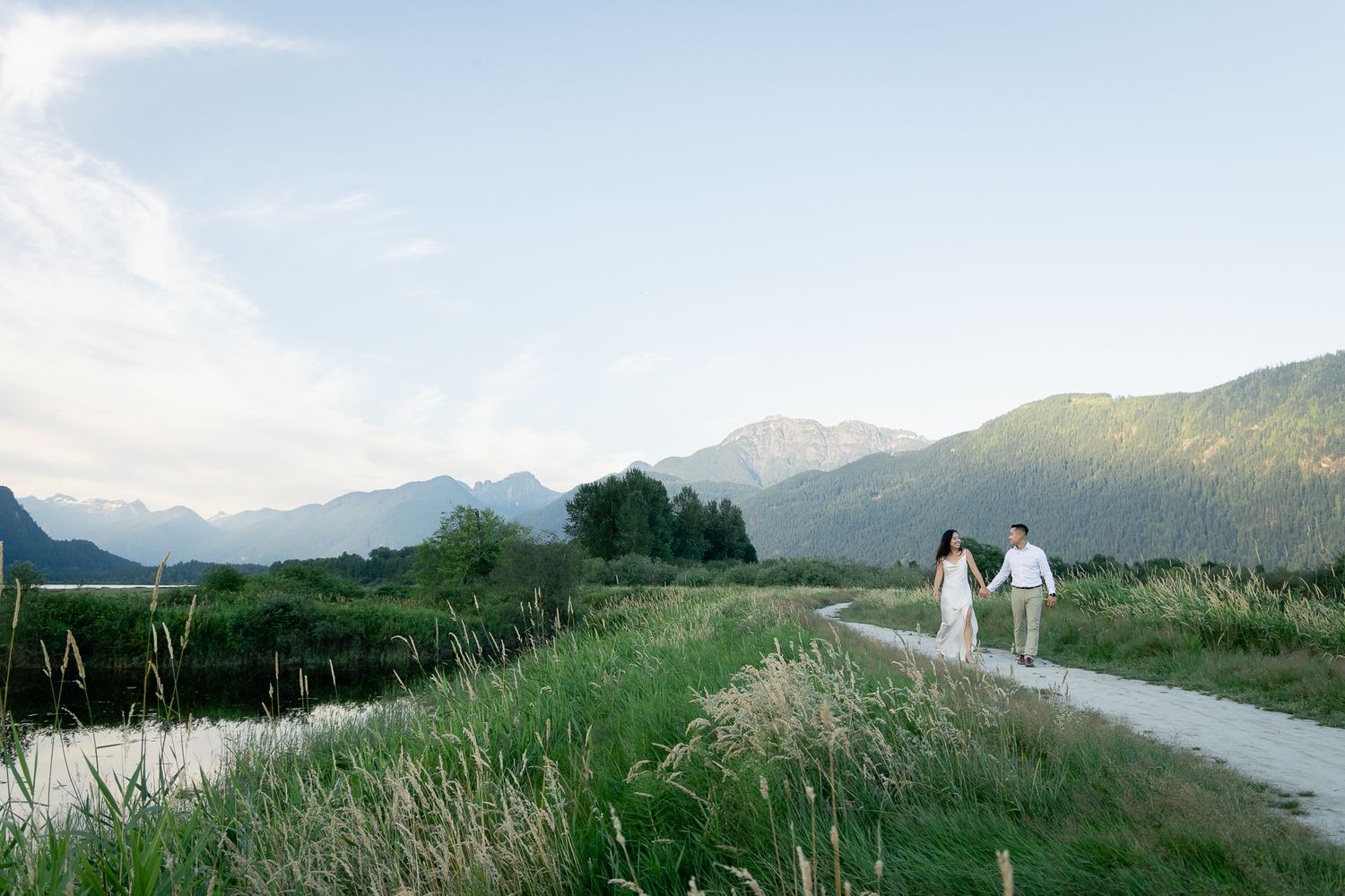 A formal engaged couple holds hands and walks down a path by Pitt Lake, lots of mountains and greenery surrounds them.
