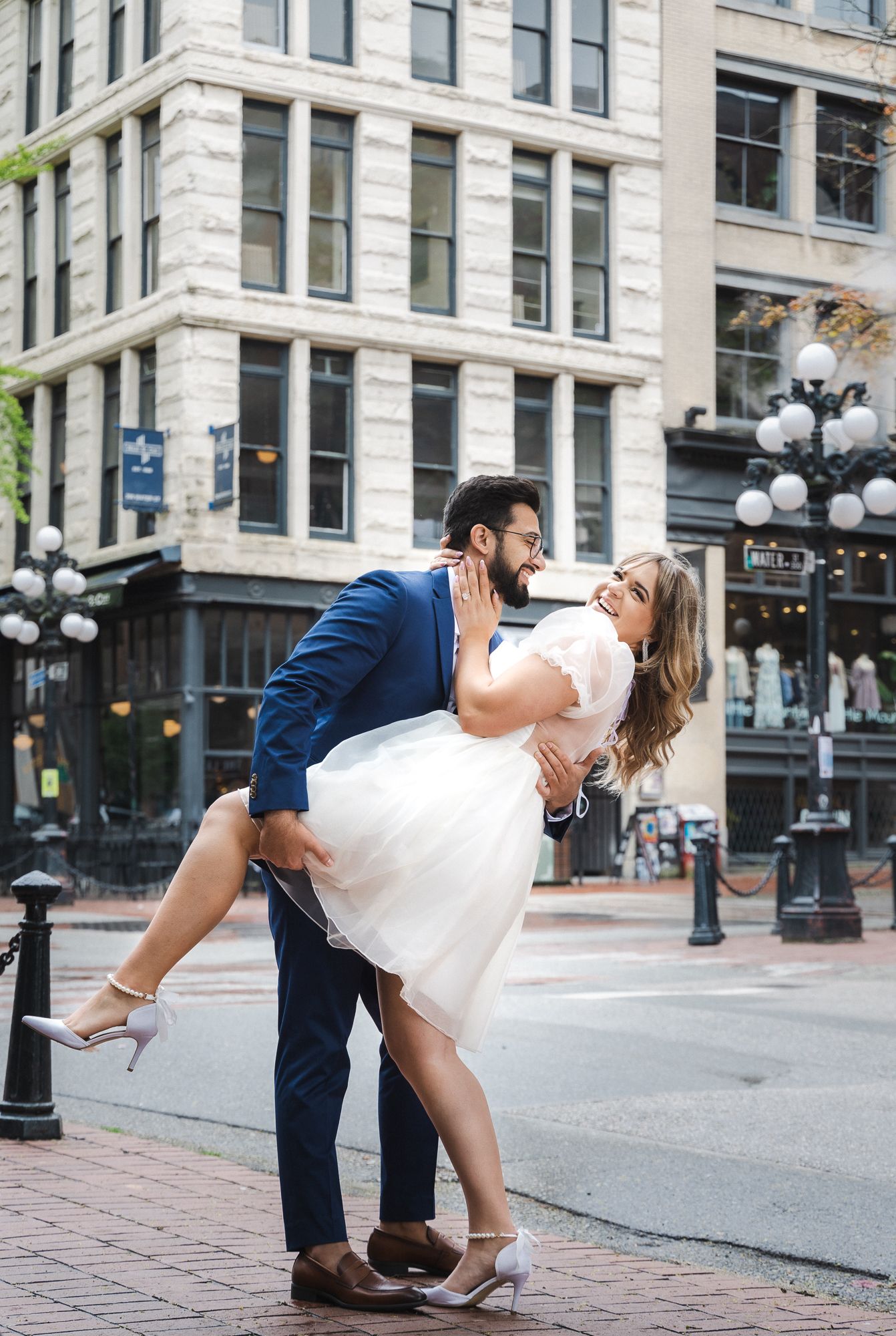 A man dips his fiancé. He looks at her lovingly while she laughs towards the camera. White dress, blue suit.