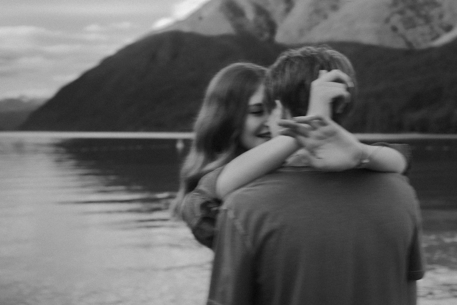 A woman teases her boyfriend, nearly kissing him, smiling. There's mountains and a lake behind.