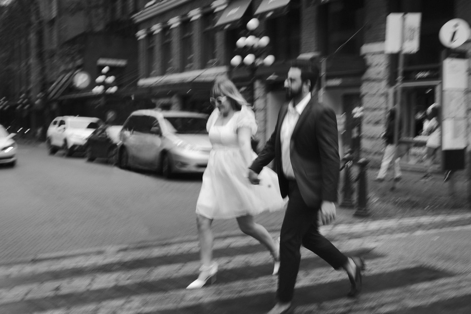 An engaged couple walks across the street in Gastown, Vancouver. She wears a white dress, he wears a black suit.