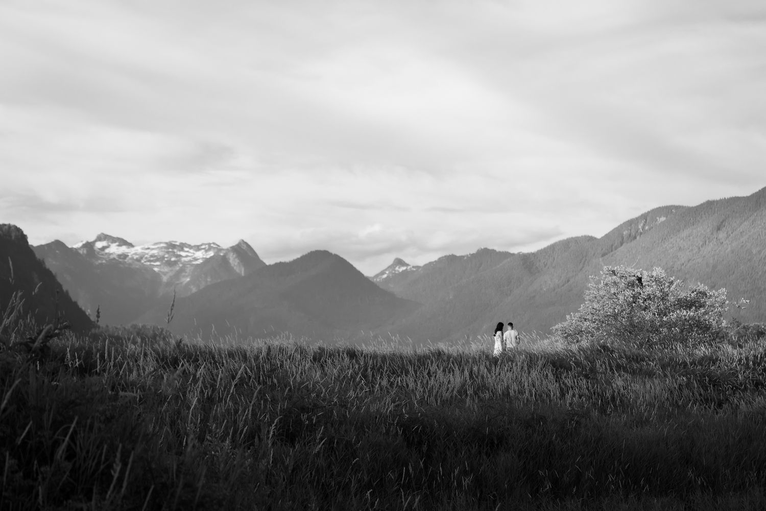 A man and woman walk together in the distance, the mountains behind them. They both wear white and look at each other.