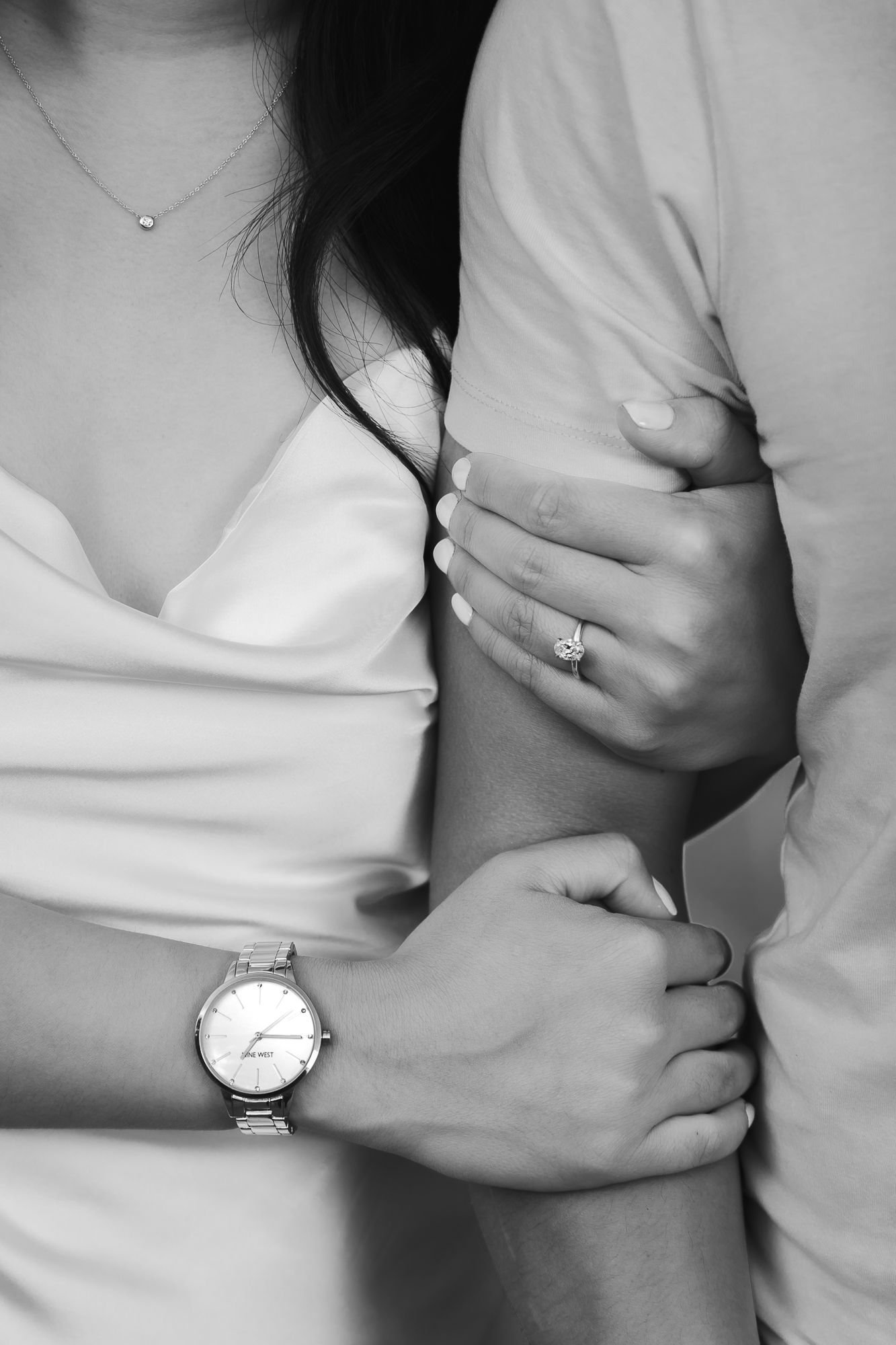 A detail shot of an engagement ring and a nice watch. The woman is holding the man's arm with both hands, showing ring.