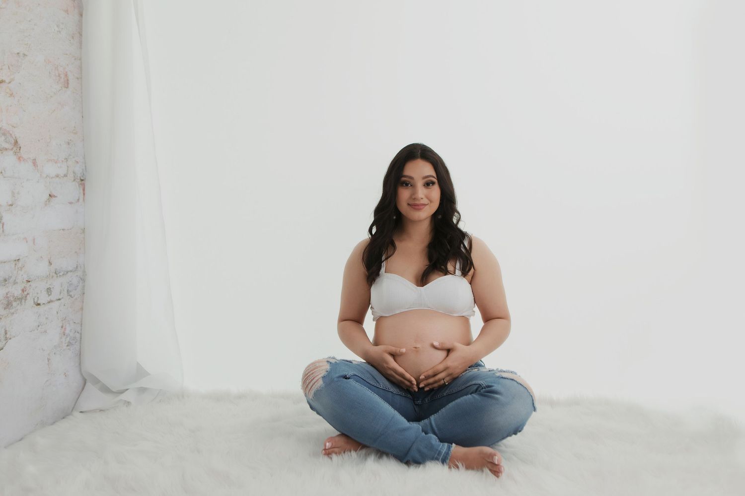 Minimalist maternity photo of woman in white top and jeans sitting cross-legged against white wall.