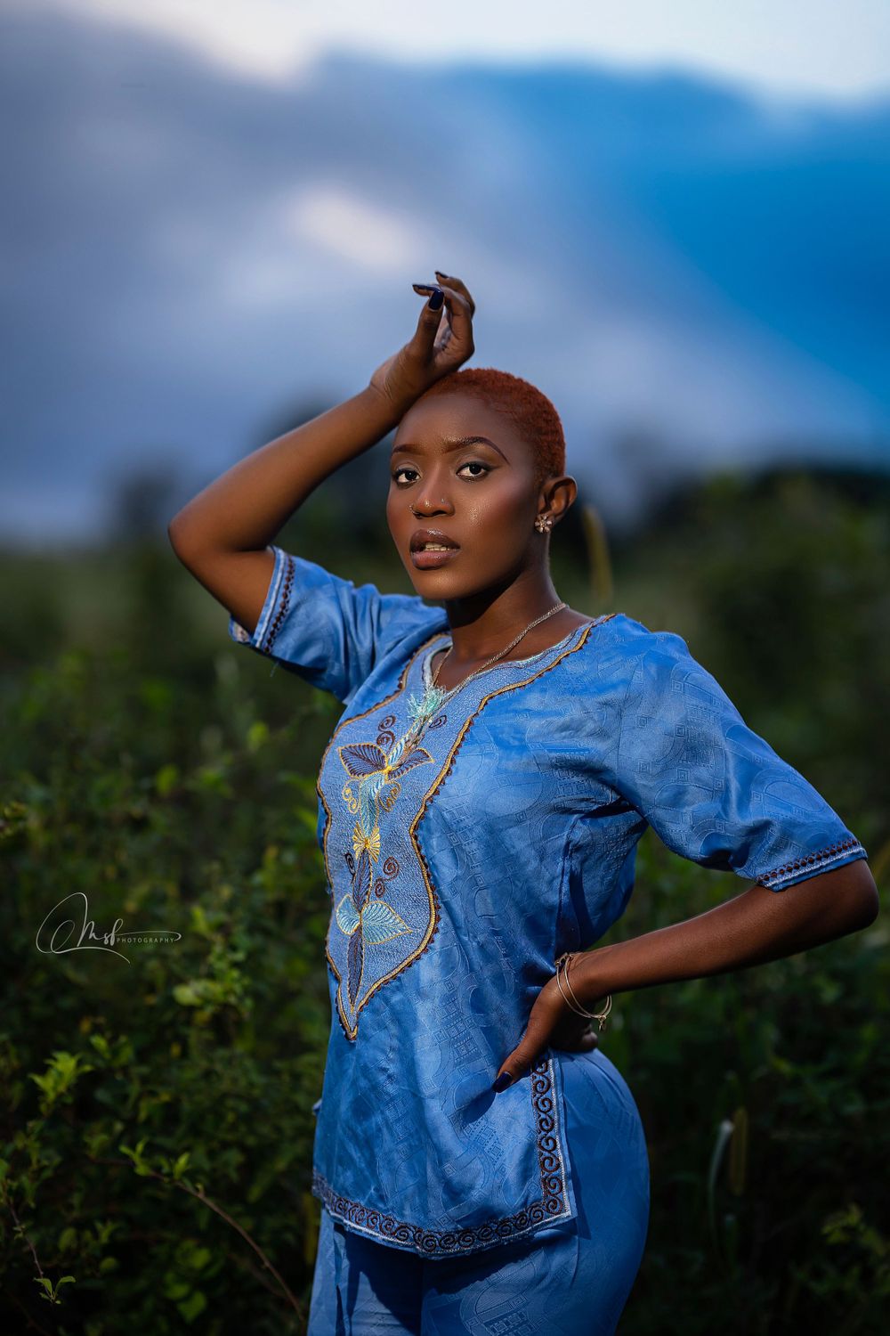 Individual in traditional blue attire poses dramatically against a dusky nature backdrop.