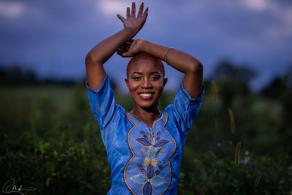 Person in a bright blue embroidered top poses joyfully with raised arms in an outdoor field at dusk.