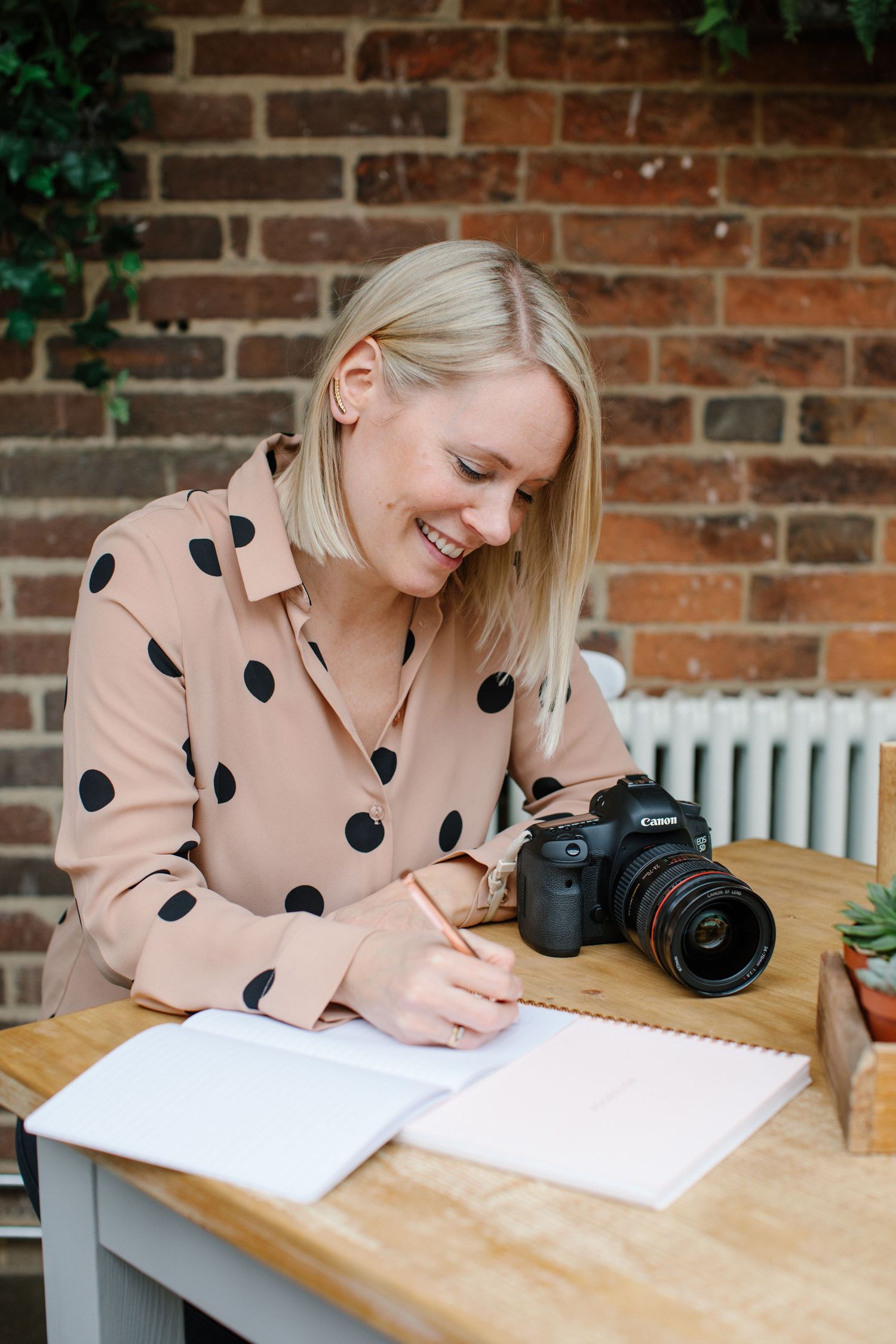 A content photographer in a polka dot blouse writes in her notebook while sitting at a wooden desk.