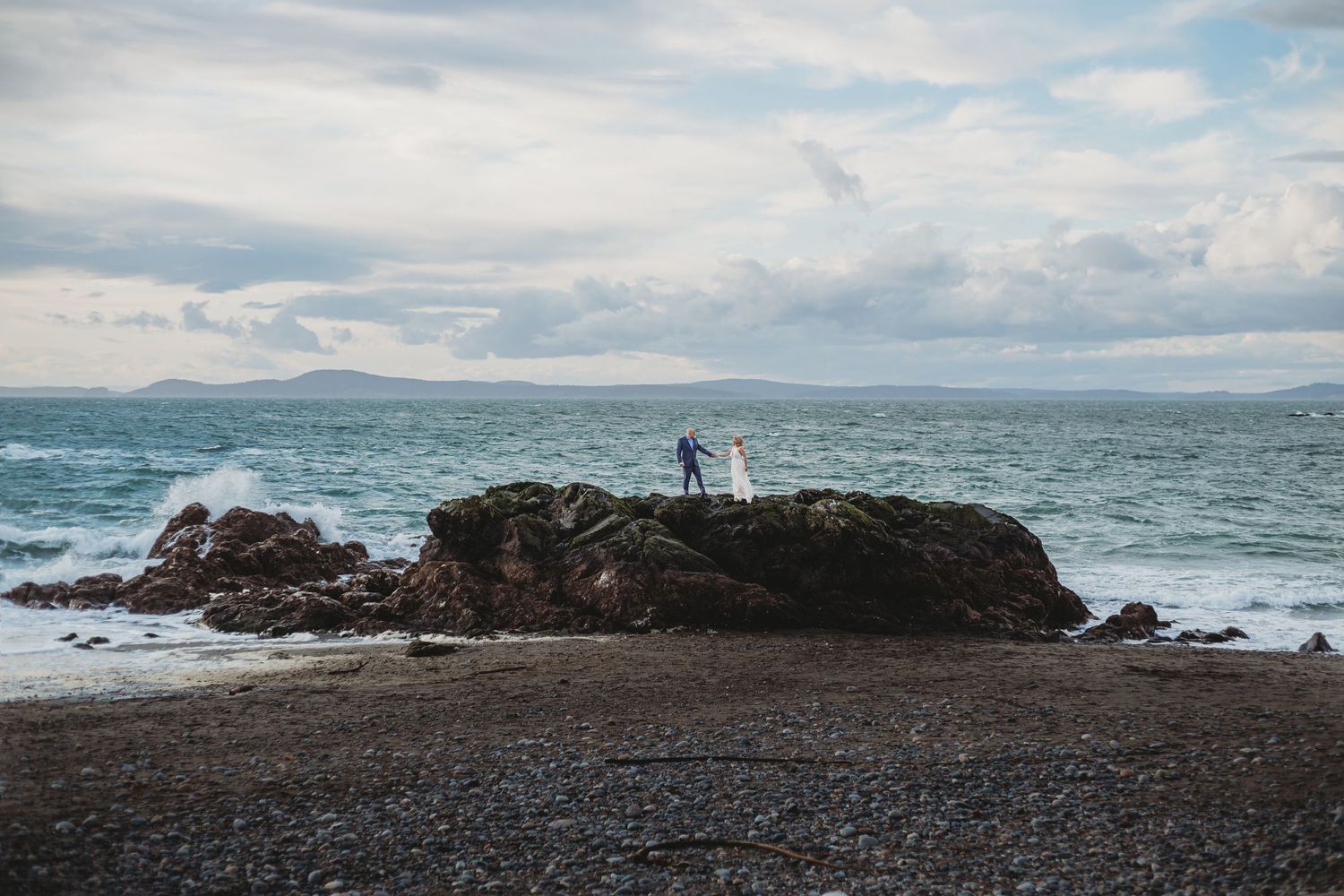 Standing atop a giant seaside rock, a bride and groom hold hands at their washington elopement