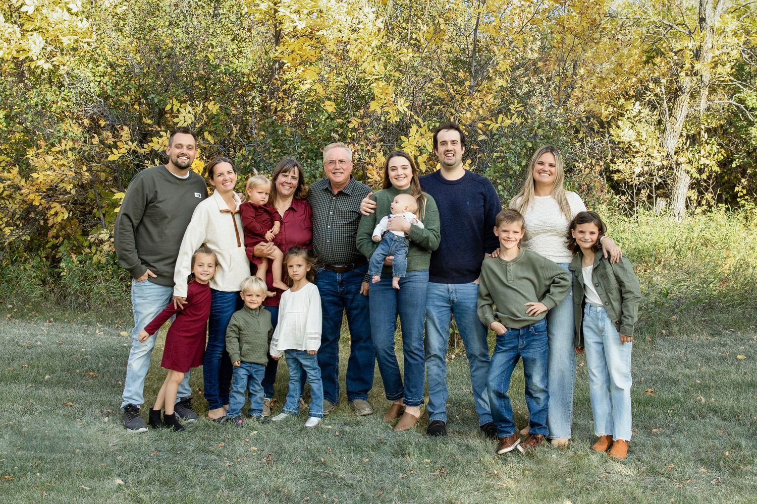 Extended family group portrait outdoors in casual clothing against autumn foliage.