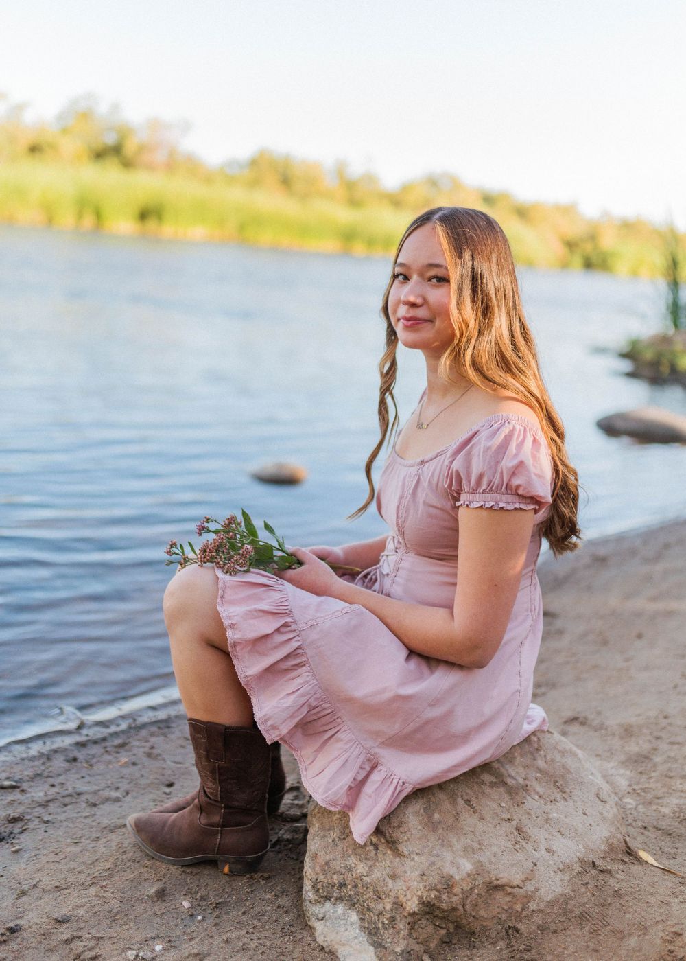 Young woman in pink dress sits by lake shore wearing brown boots in golden sunlight.