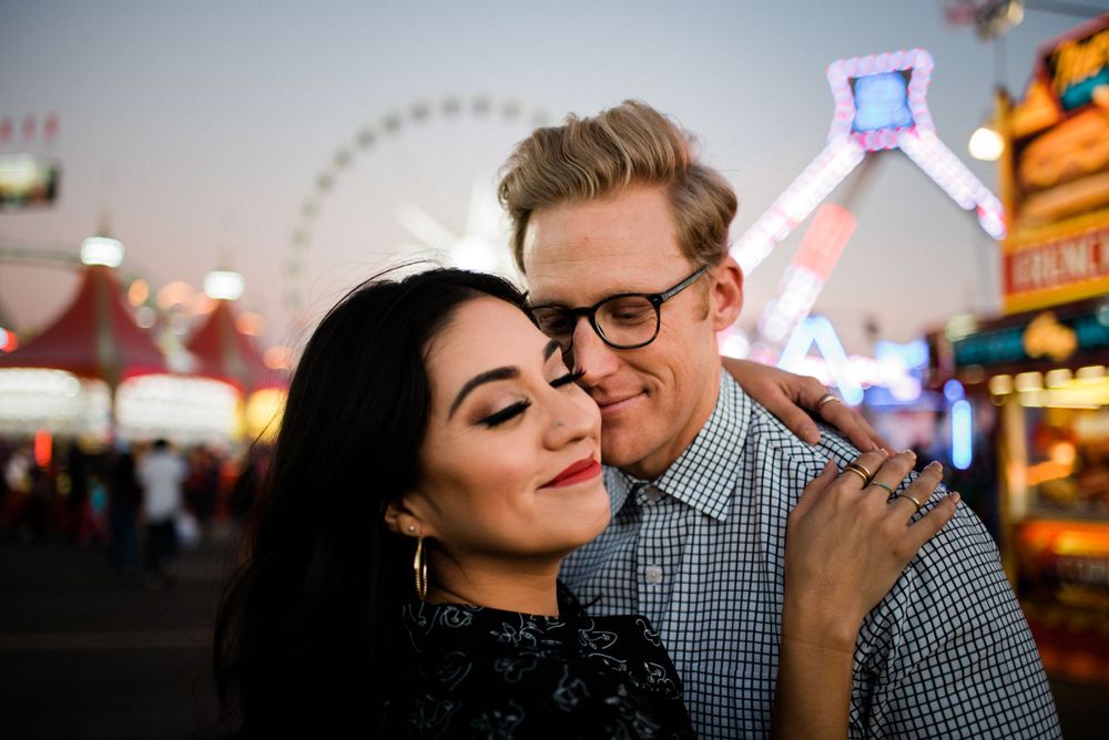 Affectionate embrace captured at dusk with carnival rides and neon lights creating romantic ambiance.