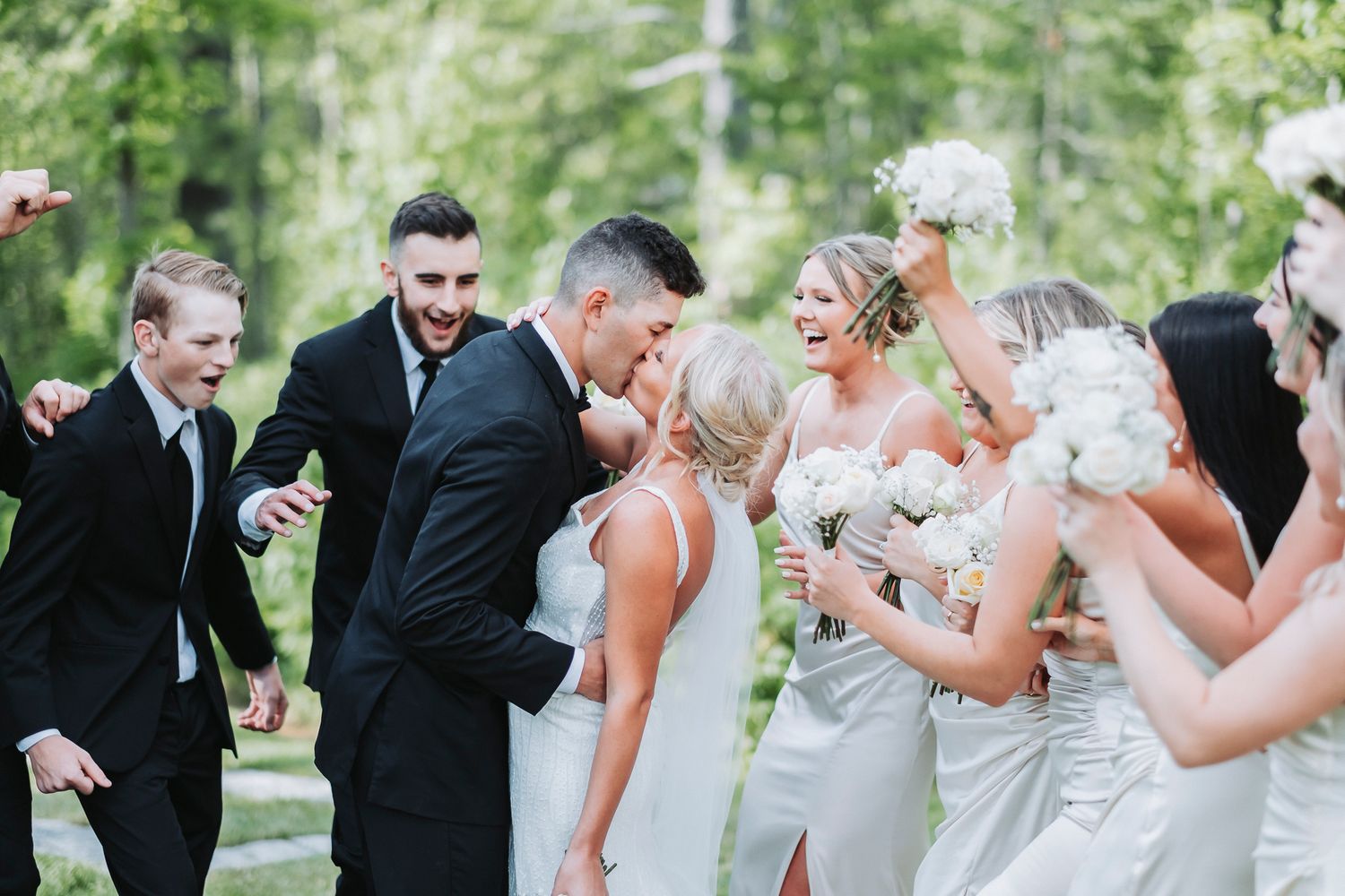 A Bride and Groom kissing, surrounded by cheering friends, against a green backdrop of trees.