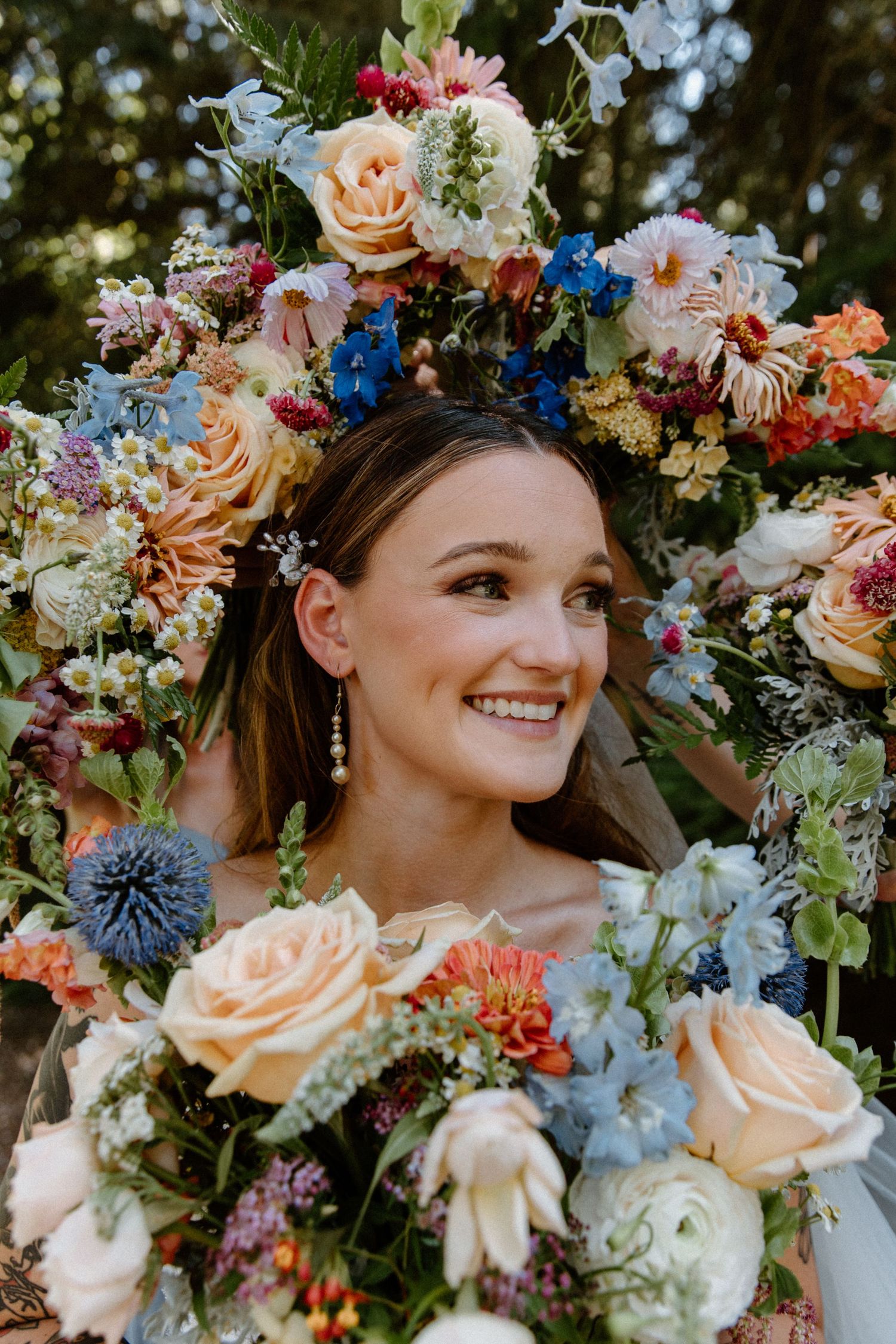 A beautiful floral crown with peach roses, blue delphiniums, and mixed wildflowers frames a joyful expression.