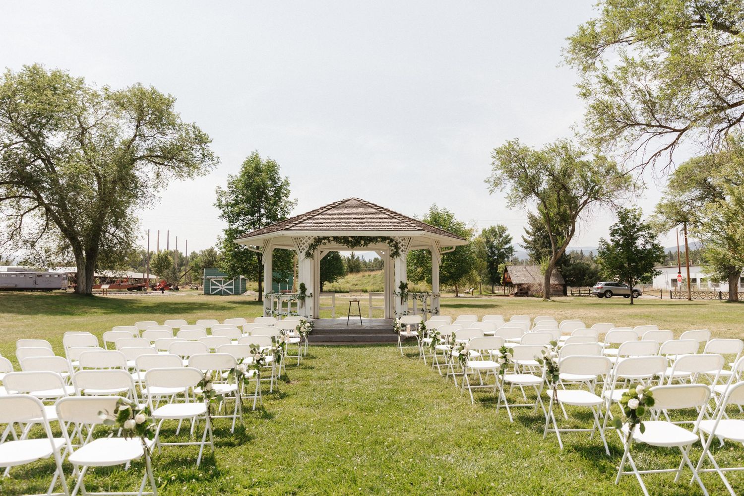 An outdoor wedding ceremony setup with white chairs leading to a gazebo surrounded by trees.