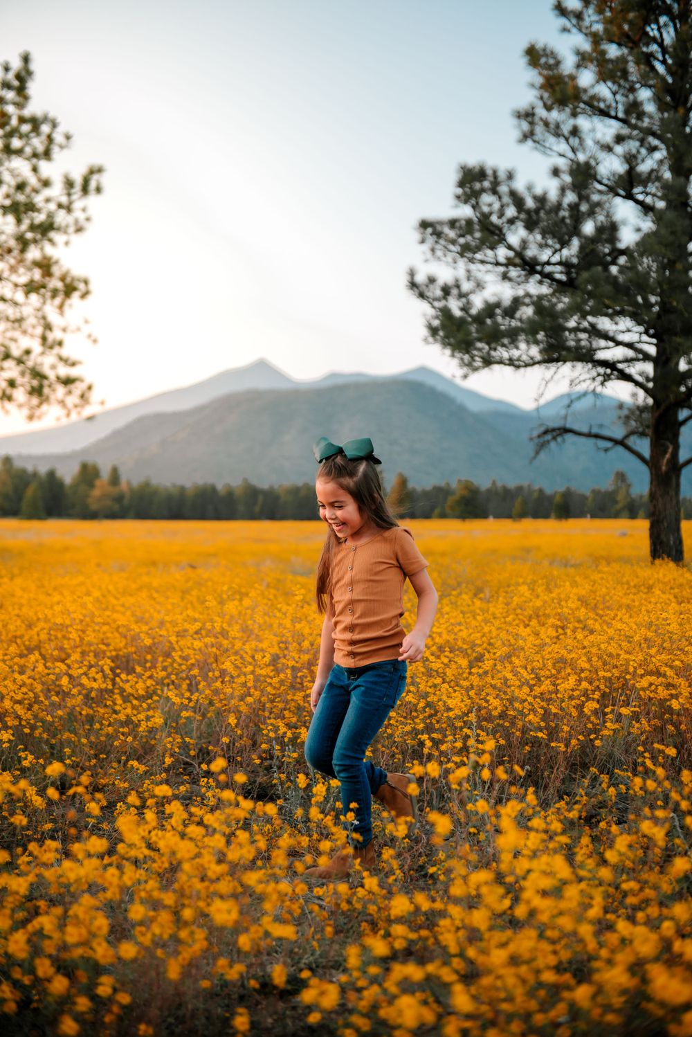 Person standing in vibrant field of yellow wildflowers with mountains in background at sunset.