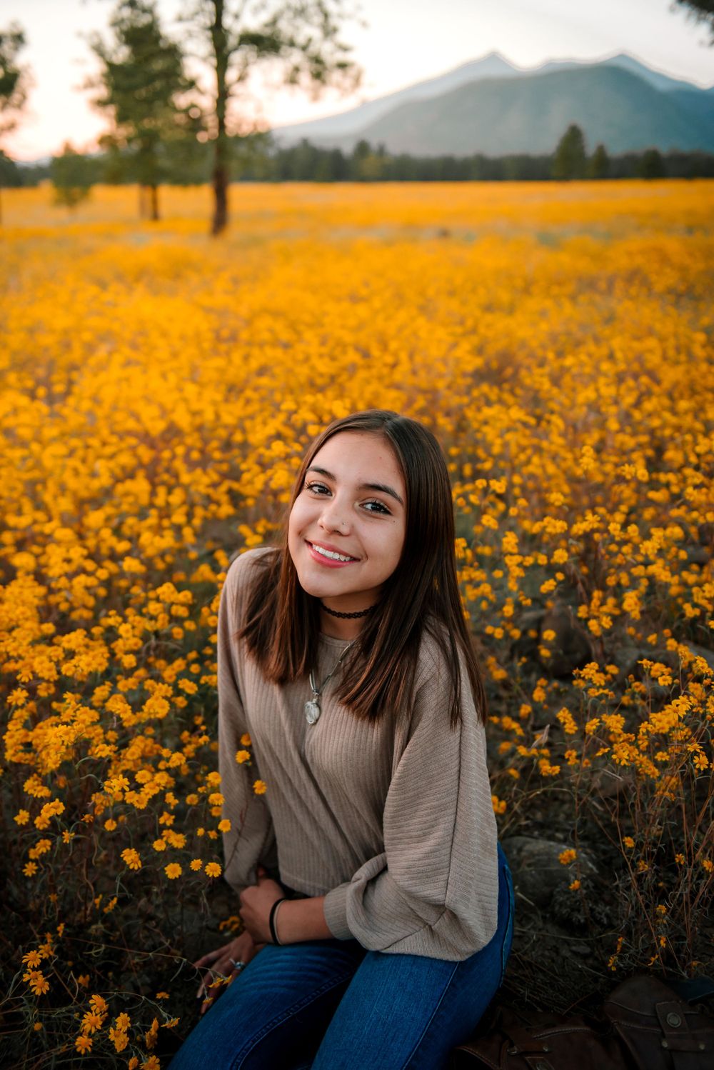 Young woman sitting in a vibrant field of yellow wildflowers with mountains in the distance.