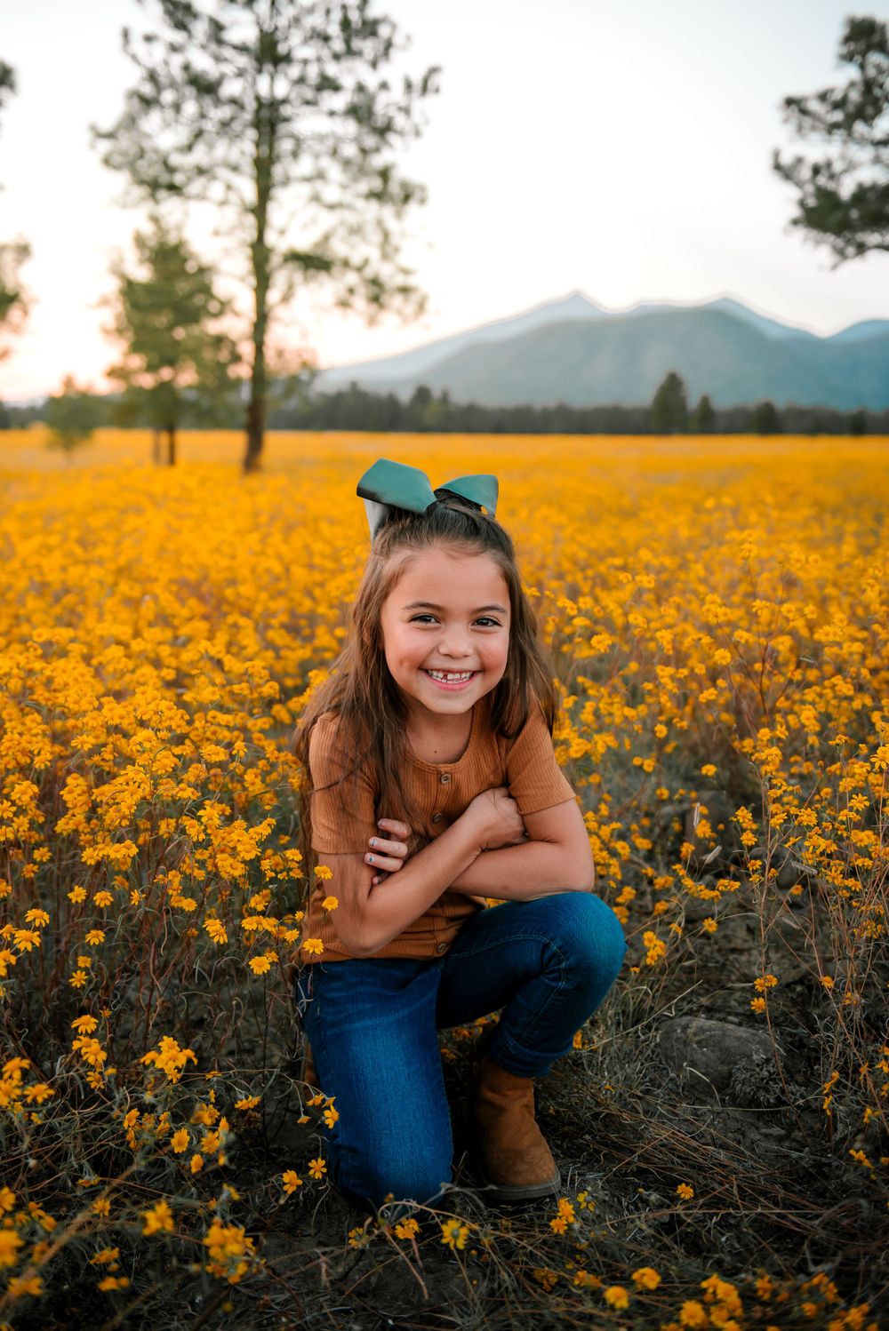 Young girl with teal bow poses in field of yellow wildflowers at sunset with mountains behind.