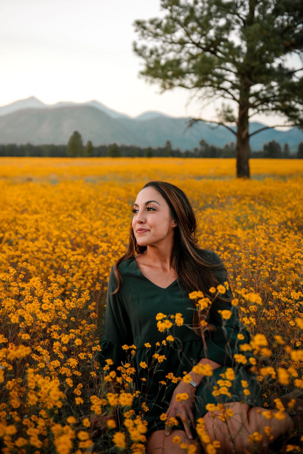 Woman in dark green sweater poses in a vast field of yellow wildflowers with mountains behind.