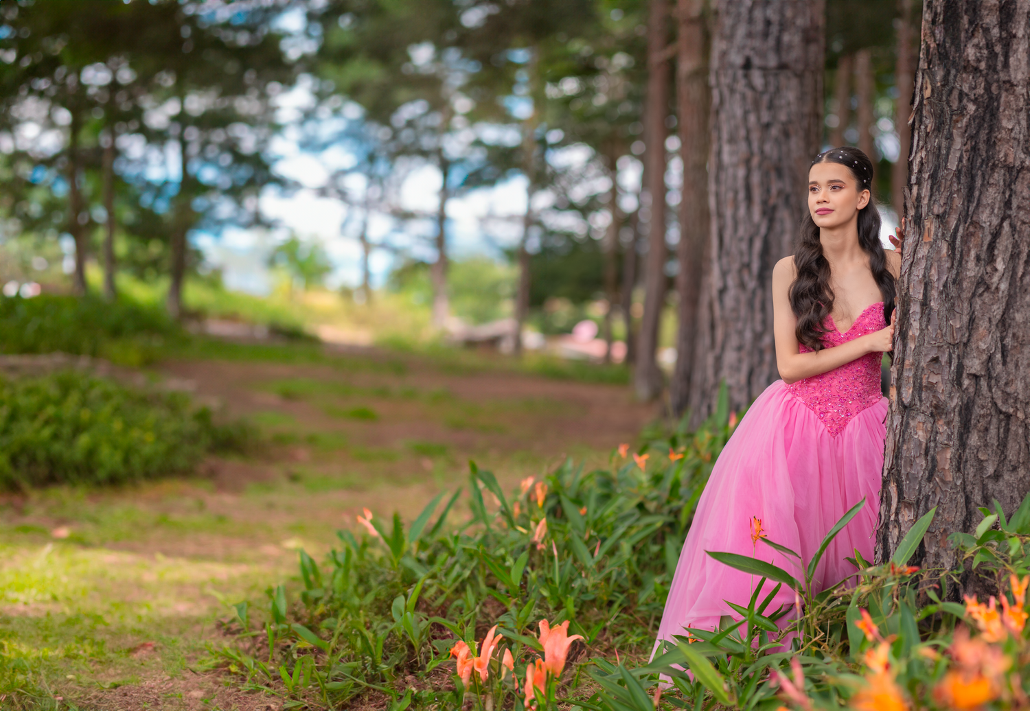 Woman in flowing pink gown poses elegantly against pine trees in a forest setting.