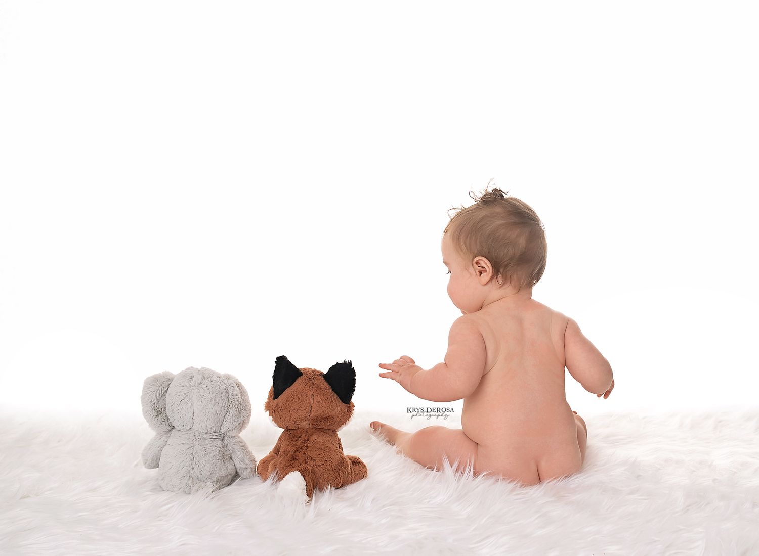 Baby sitting with plush elephant and fox toys on white fluffy backdrop.