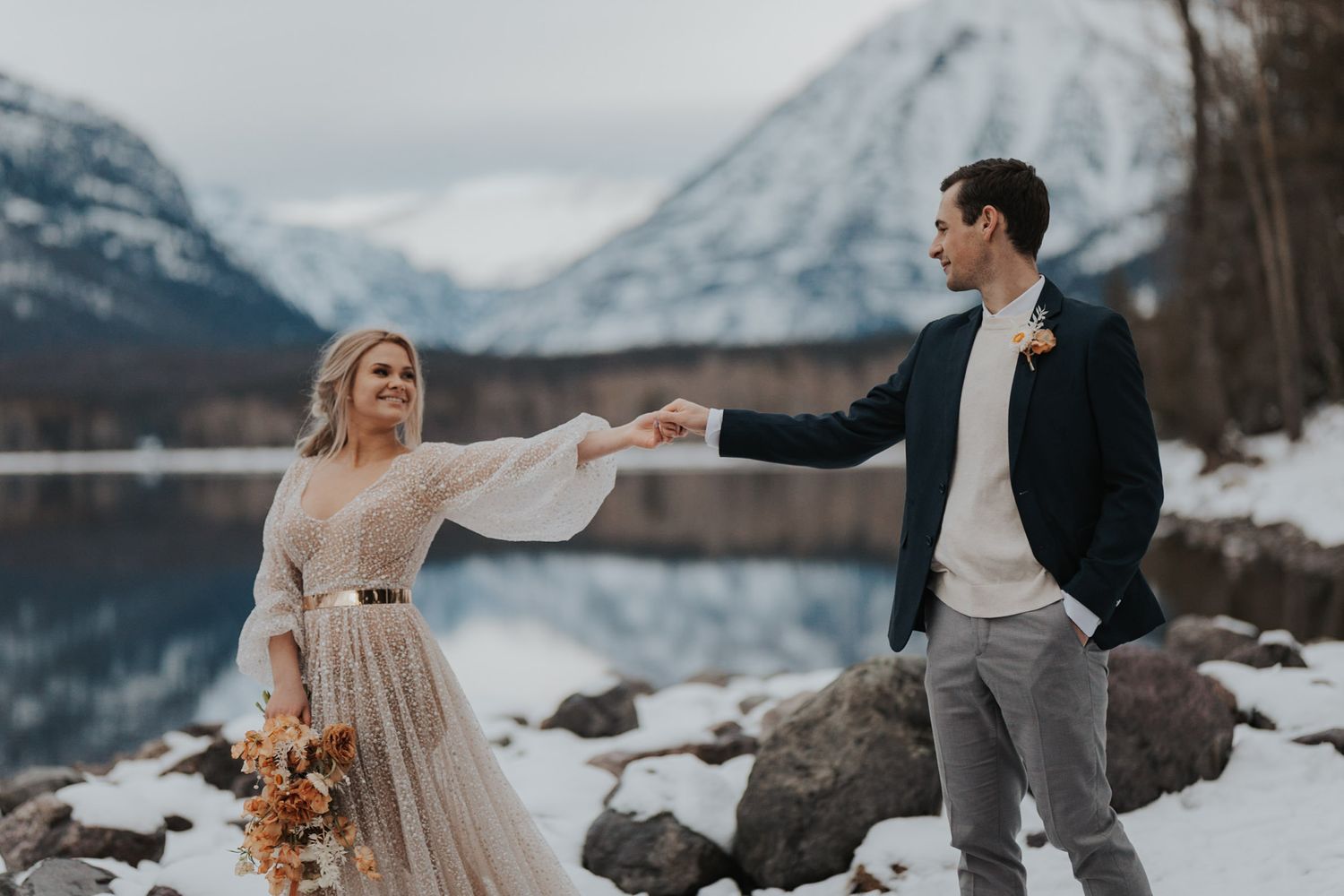 Bride and groom walk along snow covered rocks in Glacier National Park smiling at one another