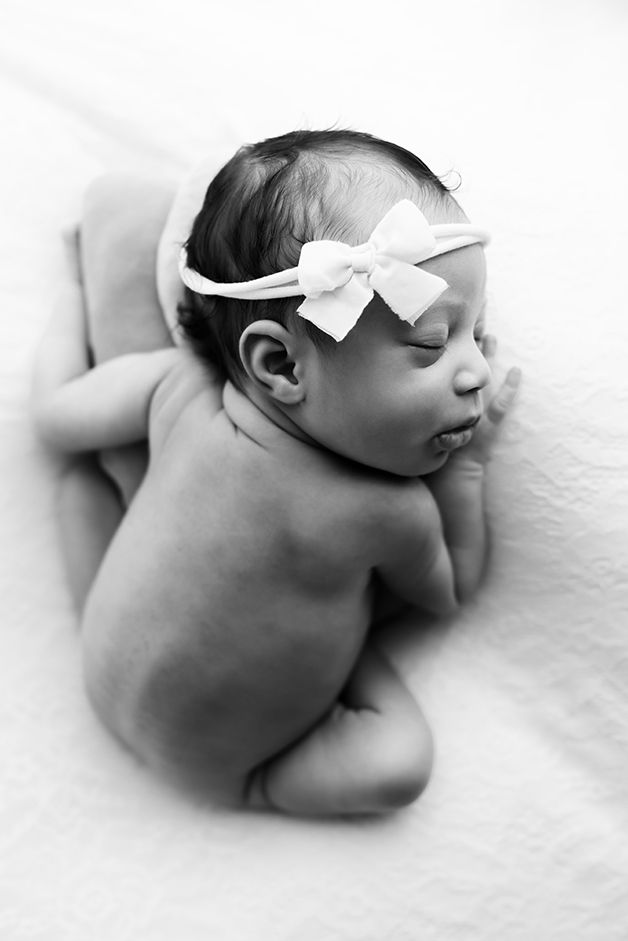 Newborn baby curled up on white blanket with white bow headband.