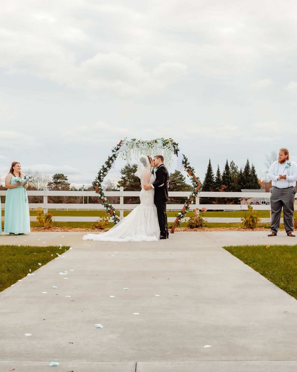 Outdoor wedding ceremony under a floral arch with white fencing and autumn scenery.