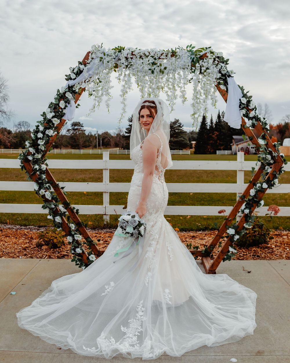 Heart-shaped floral wedding arch with cascading white flowers against white fence backdrop.