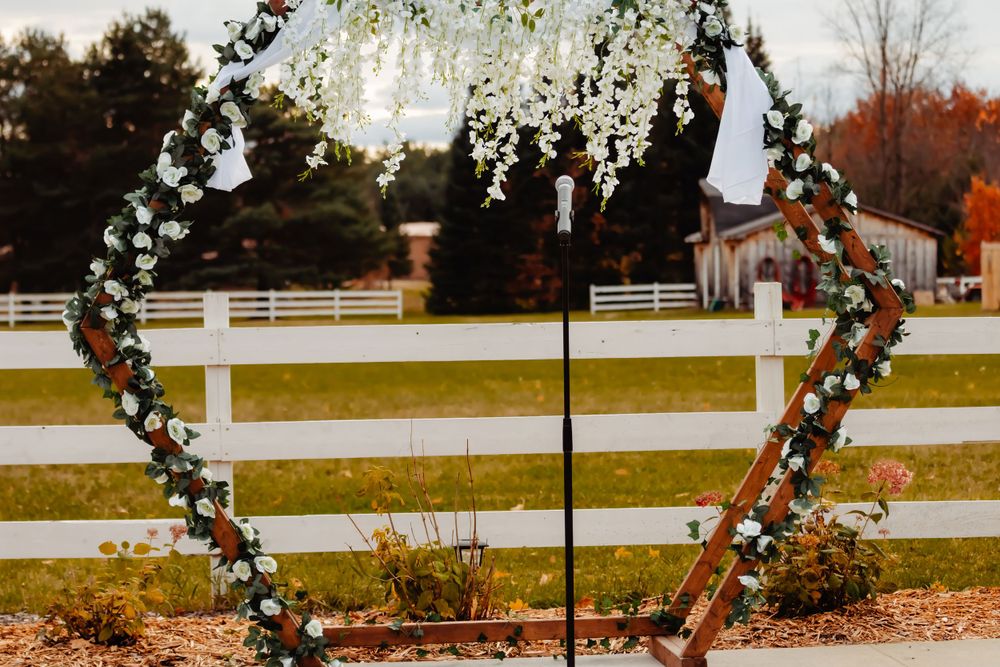Heart-shaped wedding arch decorated with white flowers and greenery against rustic farm backdrop.