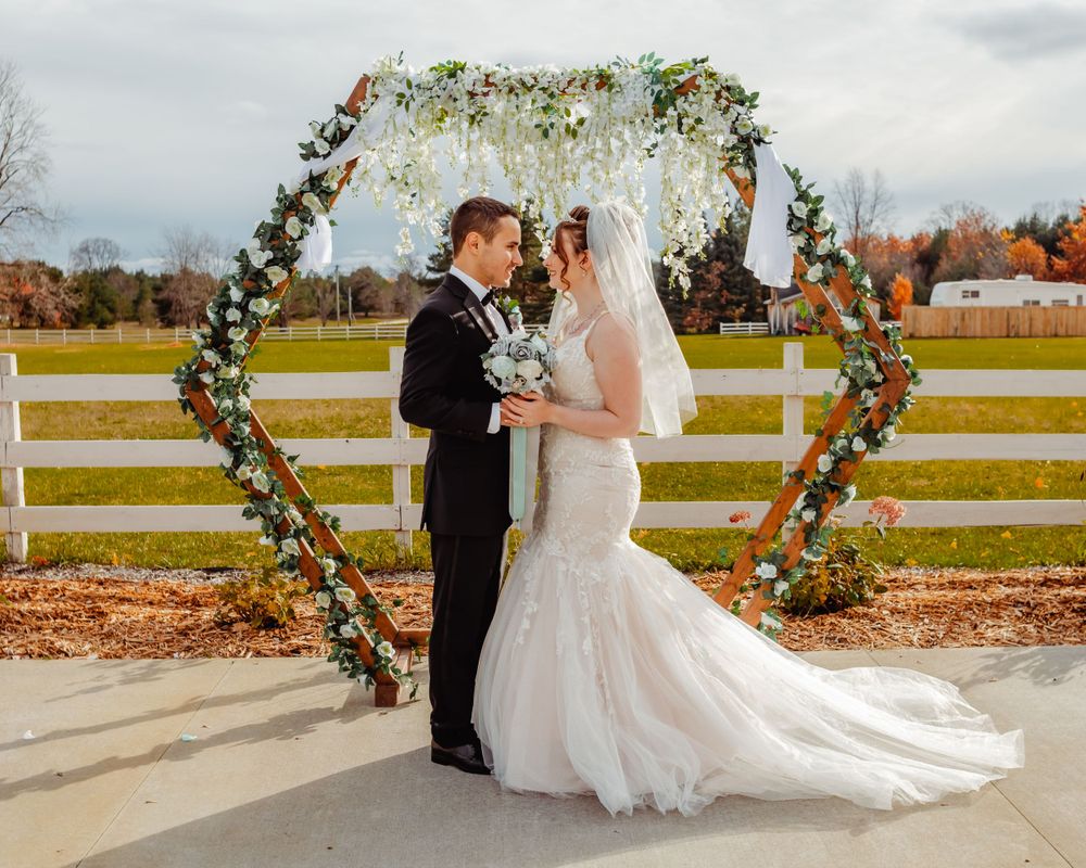 A wedding ceremony under a floral heart-shaped arch on a farm with white fences in autumn.