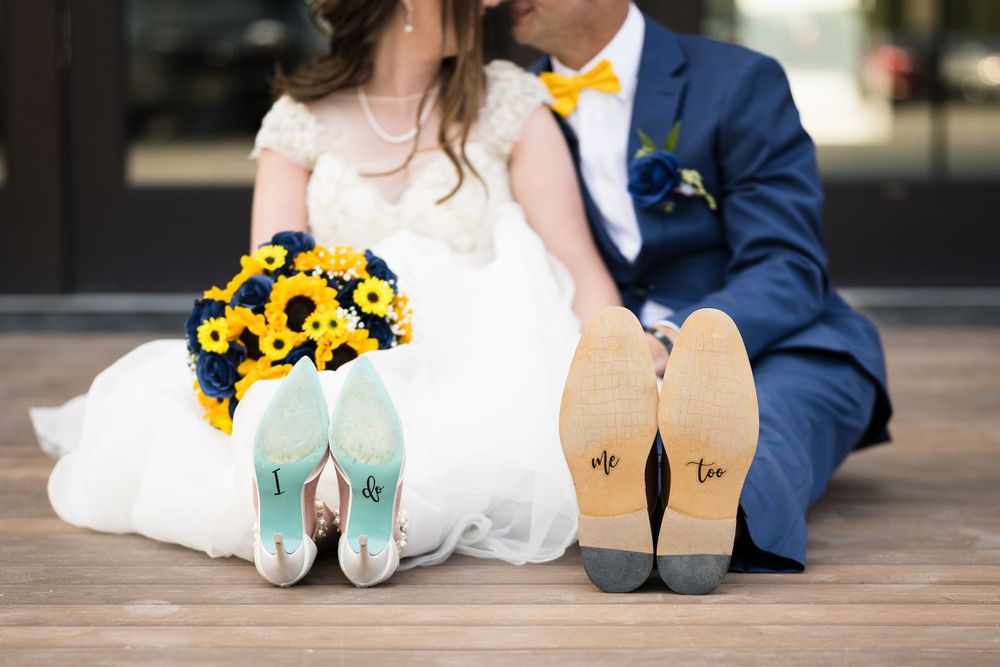 Wedding couple shows off their decorated shoe soles with sunflower bouquet on wooden deck.