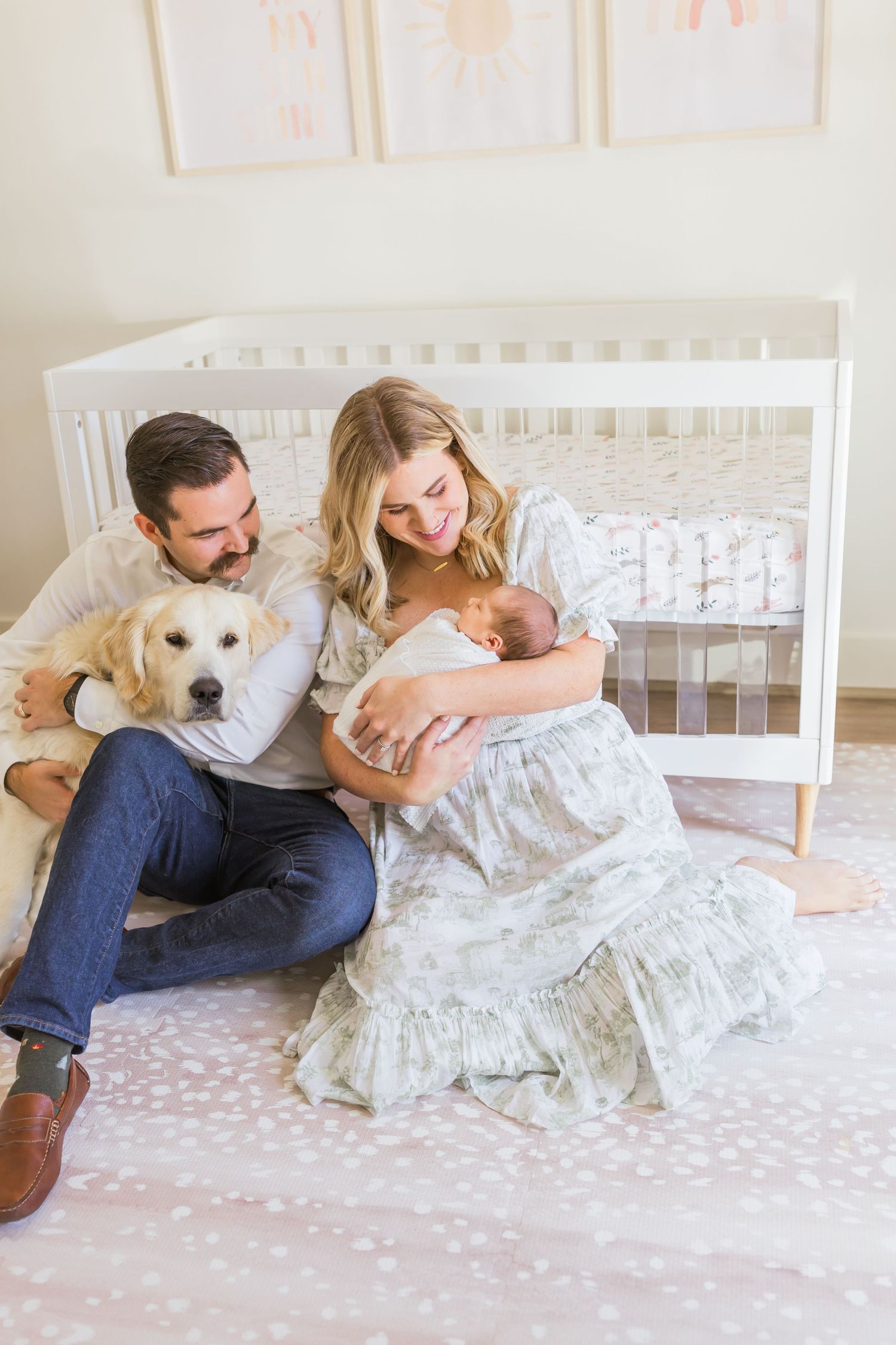 A family sitting on floor with golden retriever and newborn baby in light-filled nursery.