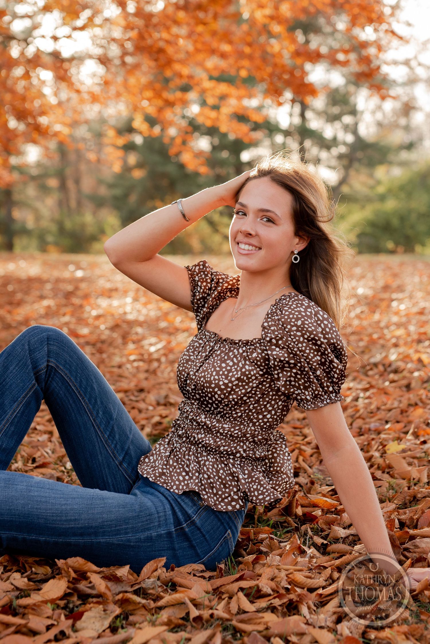 Portrait of someone in a leopard print top sitting in autumn leaves with vibrant orange foliage in background.