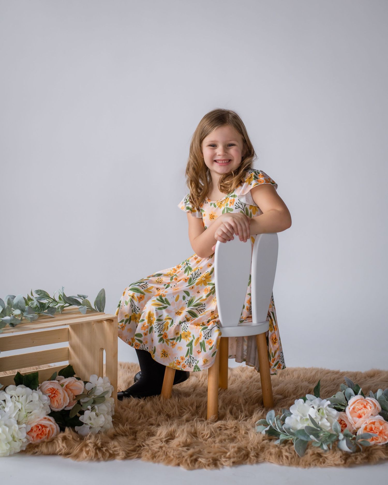 Young girl in floral dress poses on chair surrounded by flowers against grey backdrop.