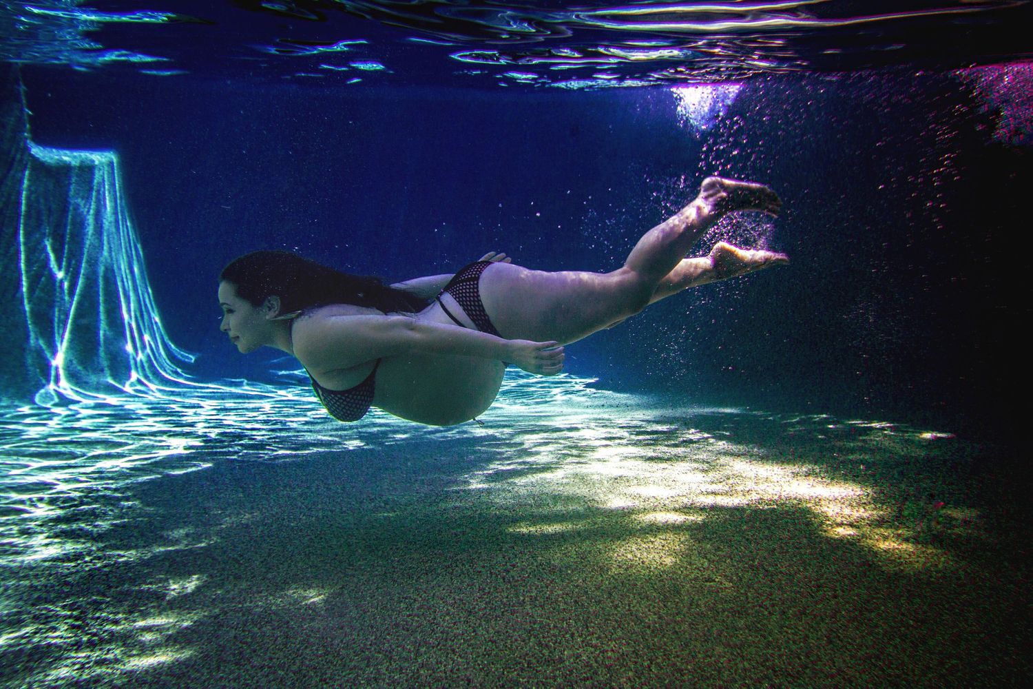Person swimming underwater at night with ethereal blue lights illuminating the dark waters in a serene aquatic scene. Underwater maternity