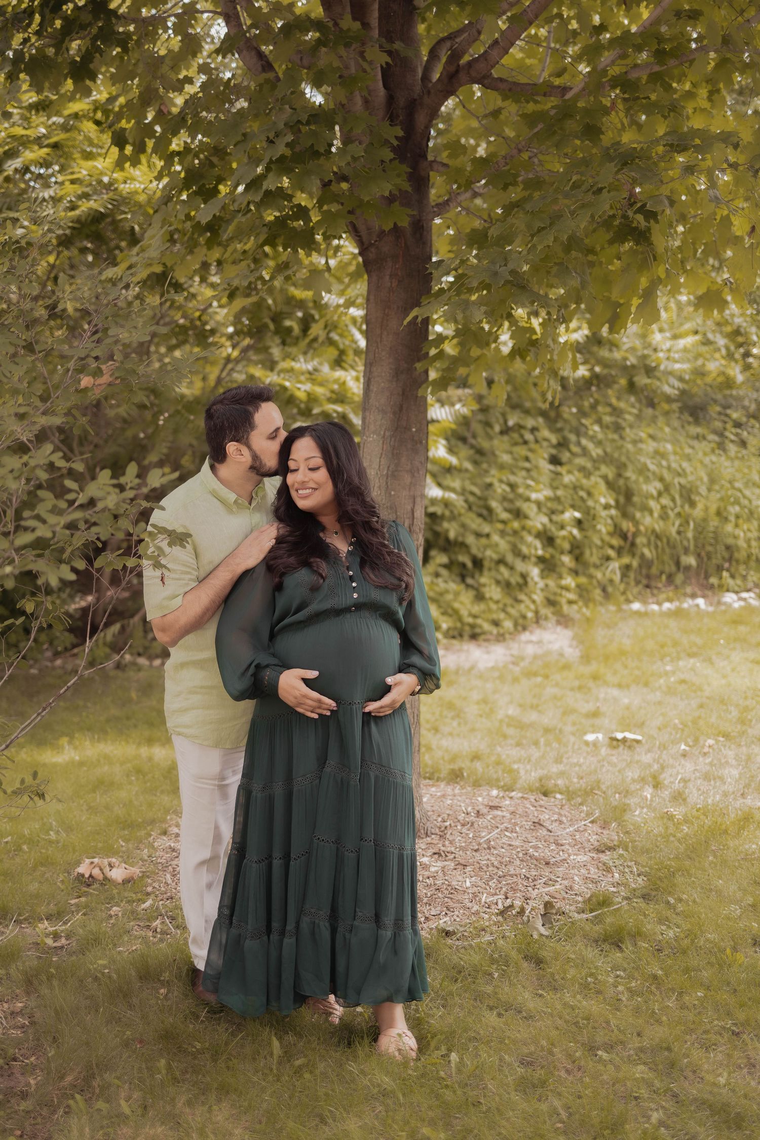 A couple shares an intimate outdoor maternity photo under a tree with the woman wearing a flowing green dress.