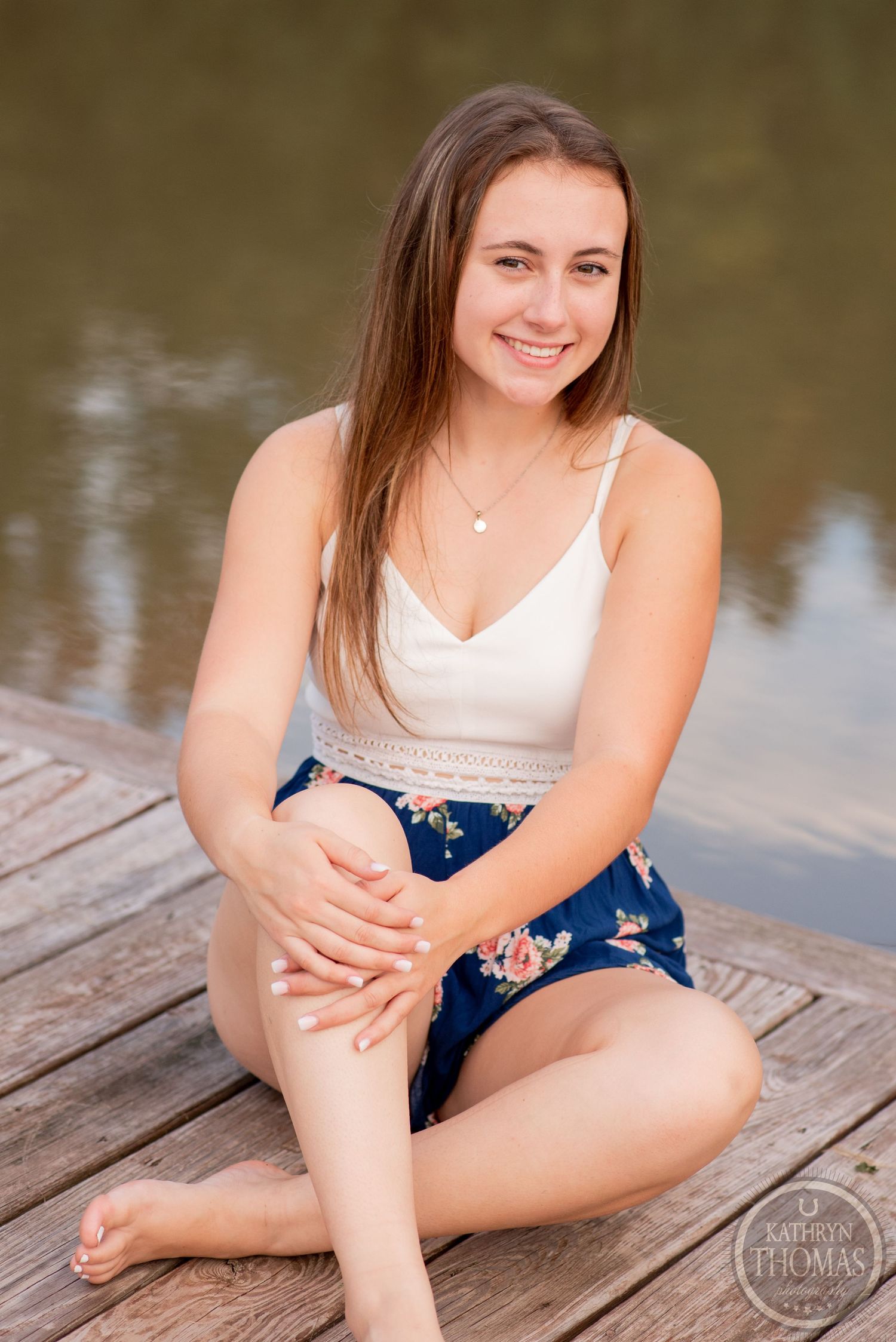A person in summer attire sits on a wooden dock with a serene lake in the background.