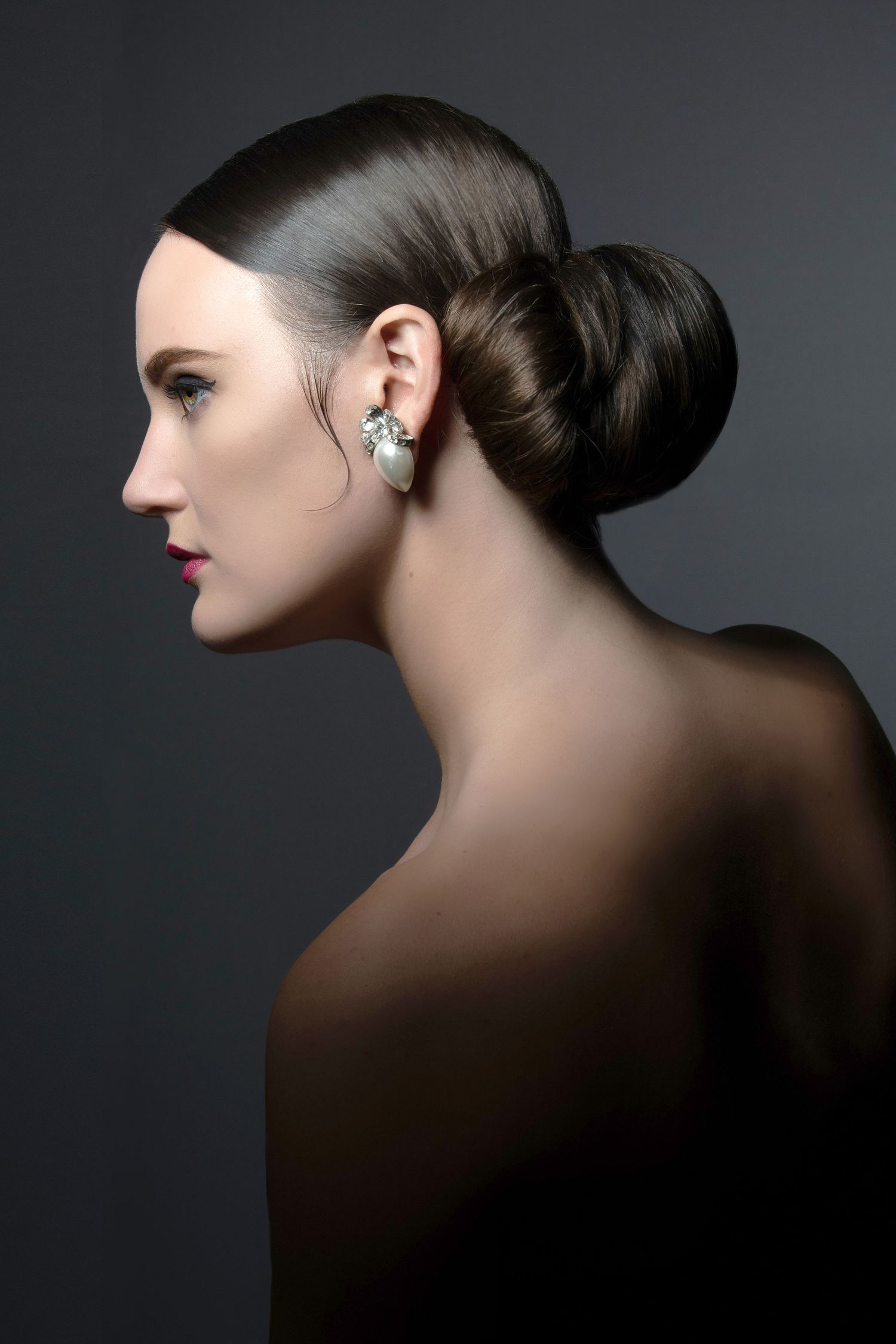 Profile view of an elegant updo hairstyle with a sparkling earring against a dark background.