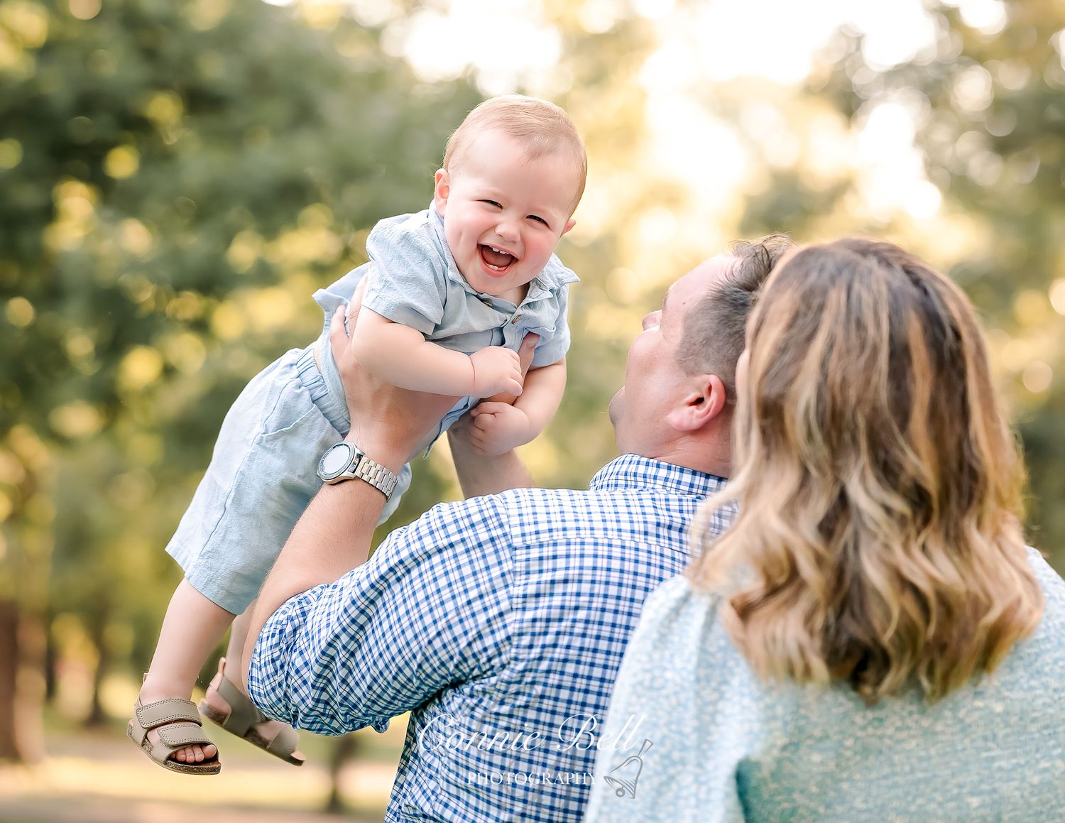 A joyful baby in light blue clothing is lifted high in the air during an outdoor family photoshoot at sunset.