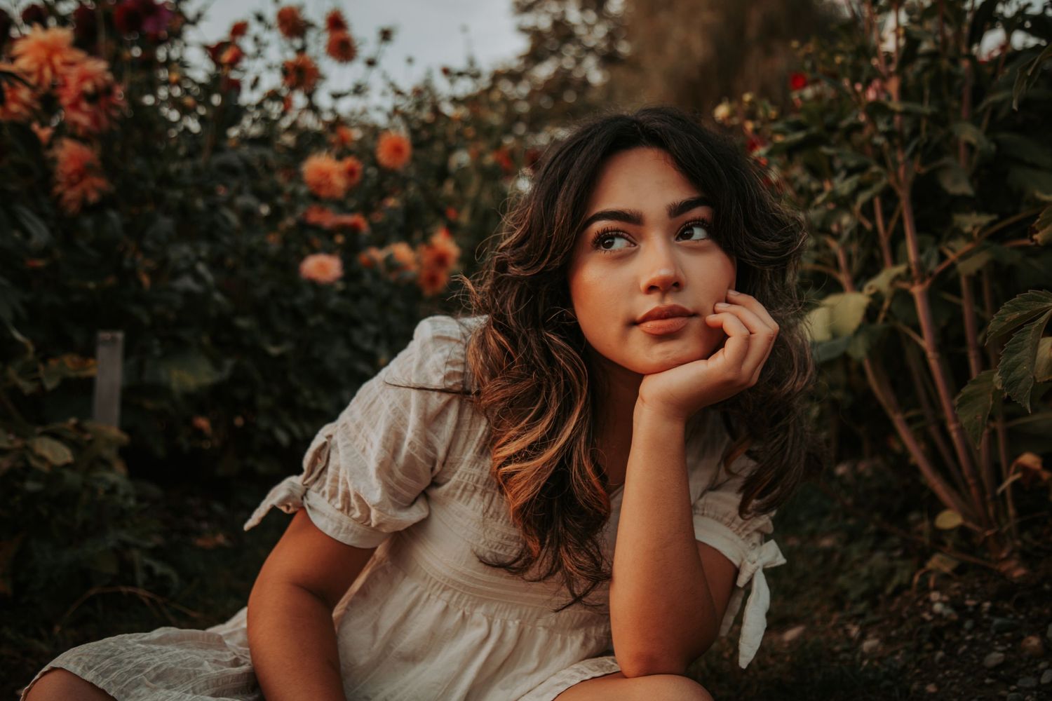Young woman with long wavy hair rests her chin on her hand while sitting among orange flowers in a garden at golden hour.