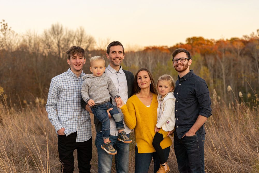 Casual outdoor family portrait in field during golden hour with autumn trees in background.